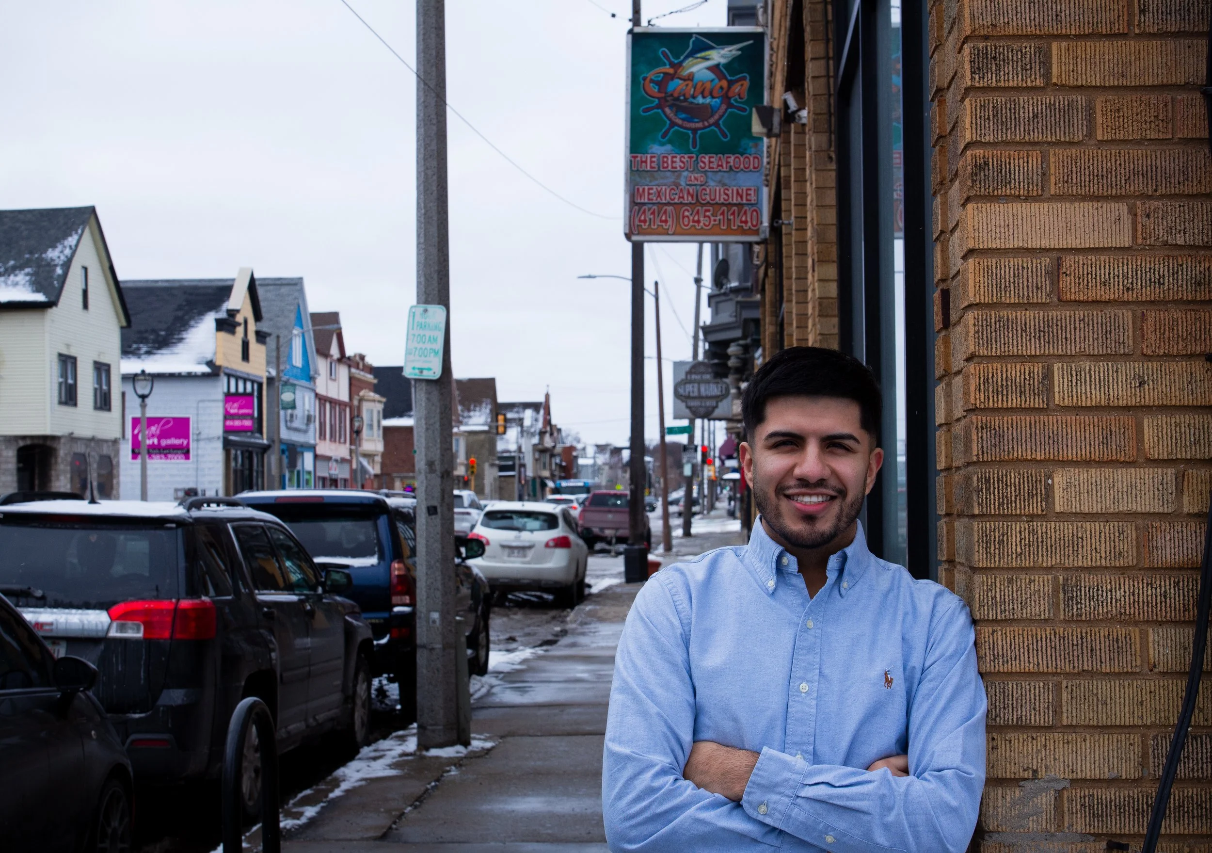 A young man with dark hair and a beard  (Ismael Luna) smiling with arms crossed, standing outdoors on a city sidewalk next to a brick building, with a street lined with cars, shops, and snow visible in the background.