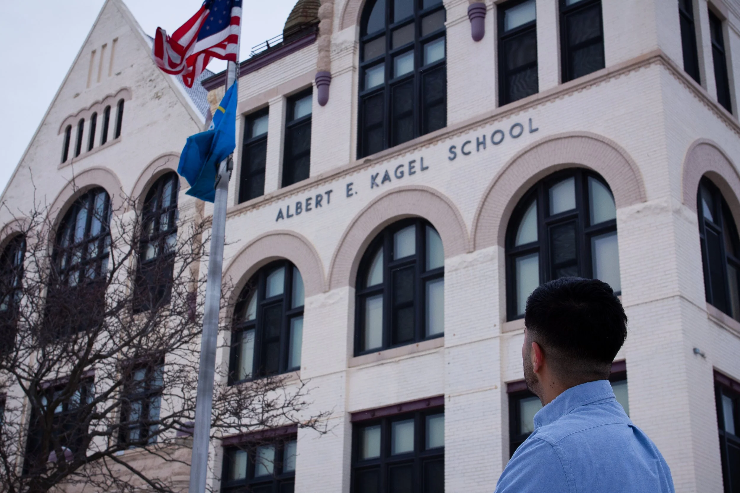 A man with dark hair (Ismael Luna) in a blue shirt looking up at a tall brick building with the sign "Albert E. Kagel School". The building has arched windows and flags, including the American flag, on a pole in front.