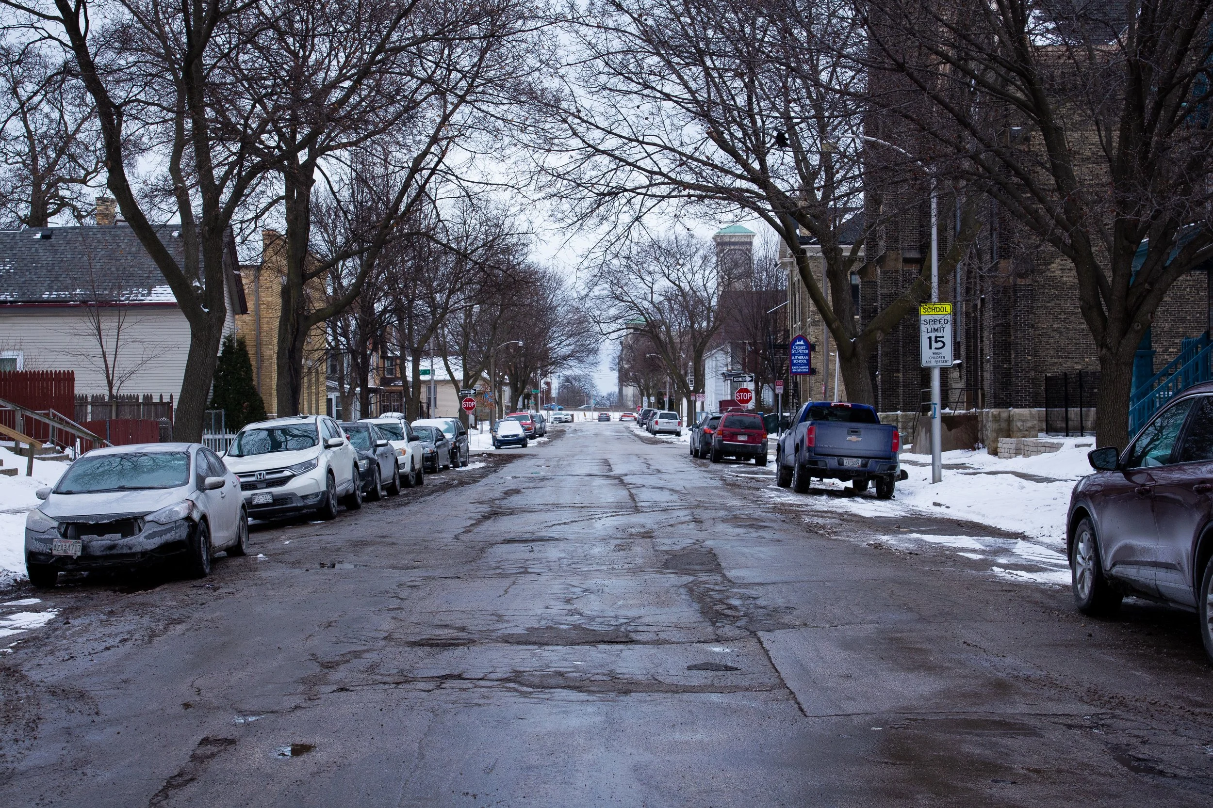A residential street in winter with snow on the ground and parked cars lining both sides of the street. The pavement is muddy and cracked, and leafless trees are overhanging the street. There are stop signs and a speed limit sign visible.