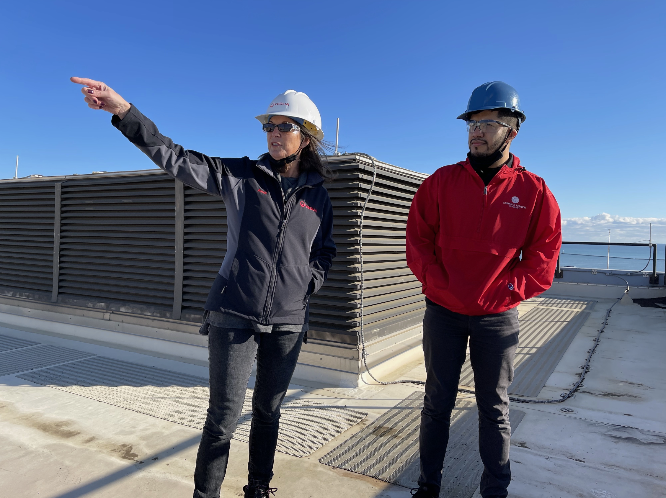 Two people (including Ismael Luna) wearing safety helmets and jackets standing on a rooftop, with one person pointing towards something in the distance against a blue sky.