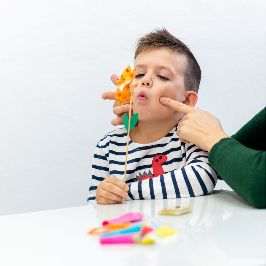 Young boy in a striped shirt is blowing a toy butterfly on a stick while a parent encourages him, with colorful toys on the table.