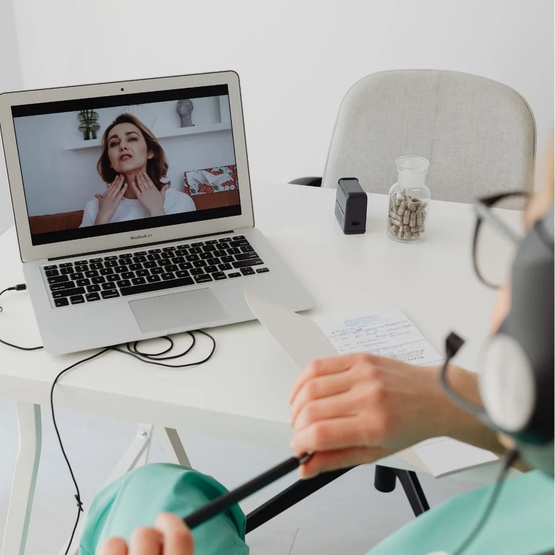 Telehealth appointment with a woman on a laptop screen showing her touching her neck, while a healthcare professional with glasses and a headset reads notes and holds a pen at a white desk.
