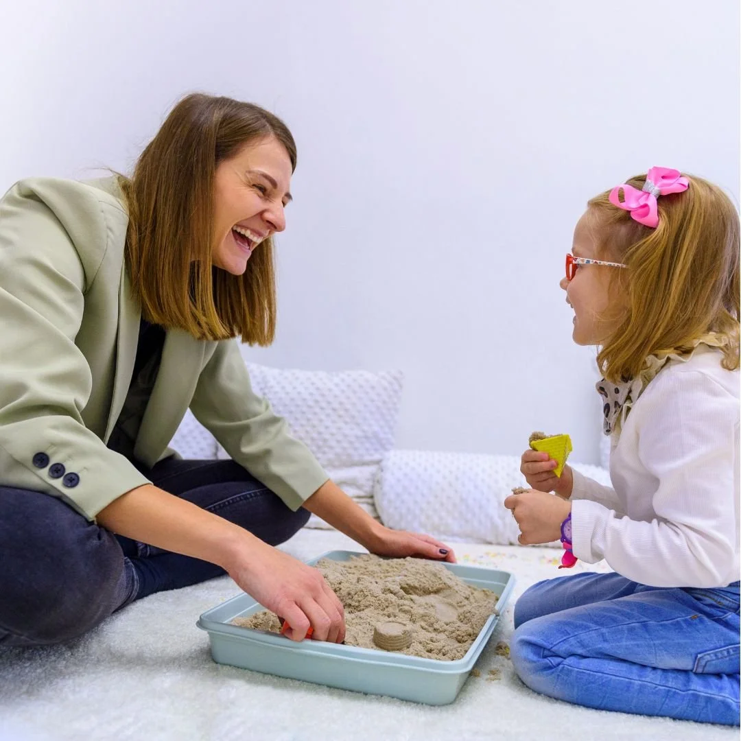 A woman and a young girl are playing together in a room with white walls. The woman is sitting on the floor, smiling and digging in a tray of sand. The girl, wearing glasses and a pink bow in her hair, is holding a small yellow shovel and a sand mold, smiling back at the woman.