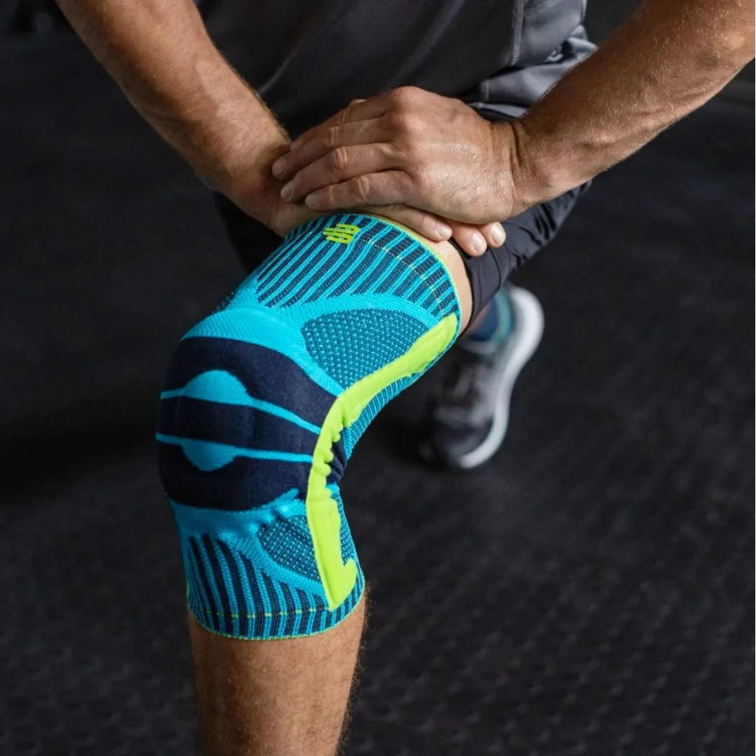 A person holding their knee, which is wrapped with a blue and neon green athletic knee brace or sleeve, in a gym or workout environment.