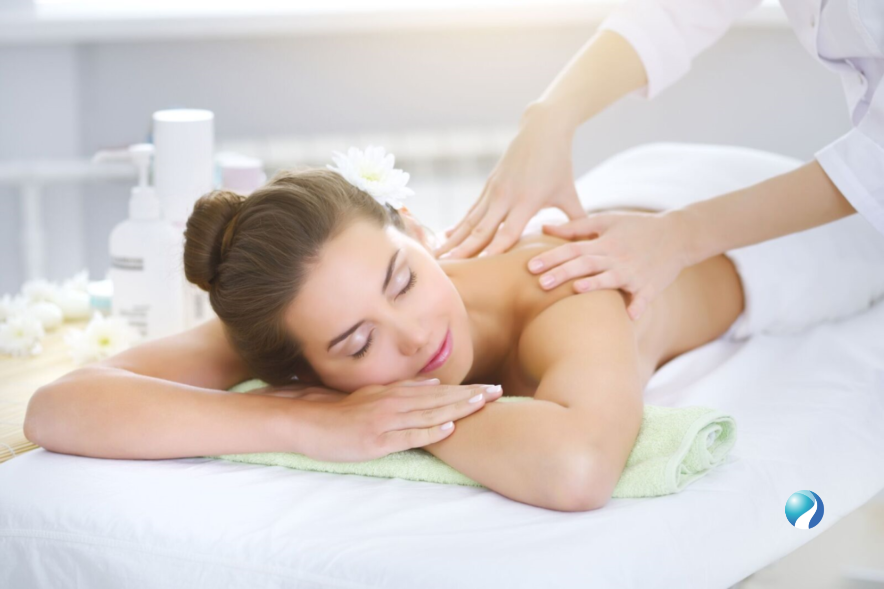 A woman receiving a massage in a spa, lying face down on a massage table with a towel under her head. The massage therapist is applying pressure to her back with both hands.
