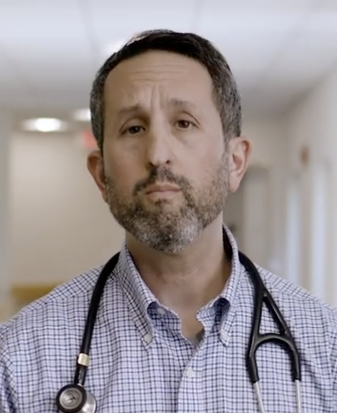A doctor with a stethoscope around his neck, standing in a hospital hallway, looking slightly skeptical or confused.