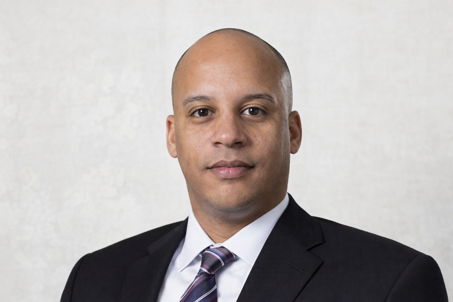 Headshot of a man in a black suit, white shirt, and striped tie, looking directly at the camera against a plain light-colored background.