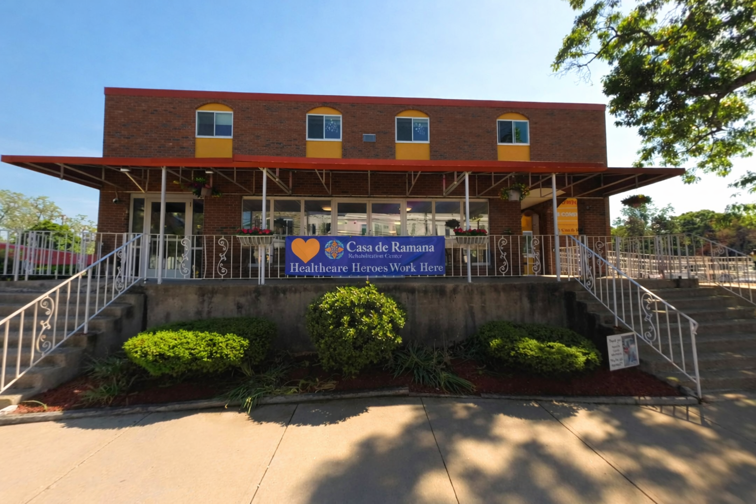 Front view of a two-story brick building with a sign that reads 'Casa de Ramana Rehabilitation Center Healthcare Heroes Work Here,' featuring windows with yellow accents on the upper floor, a red metal awning, white railings, and landscaped bushes in front.