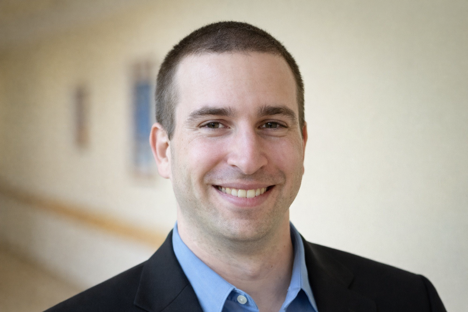 Headshot of a smiling man in a suit with a light blue shirt, indoors with a blurred hallway background.