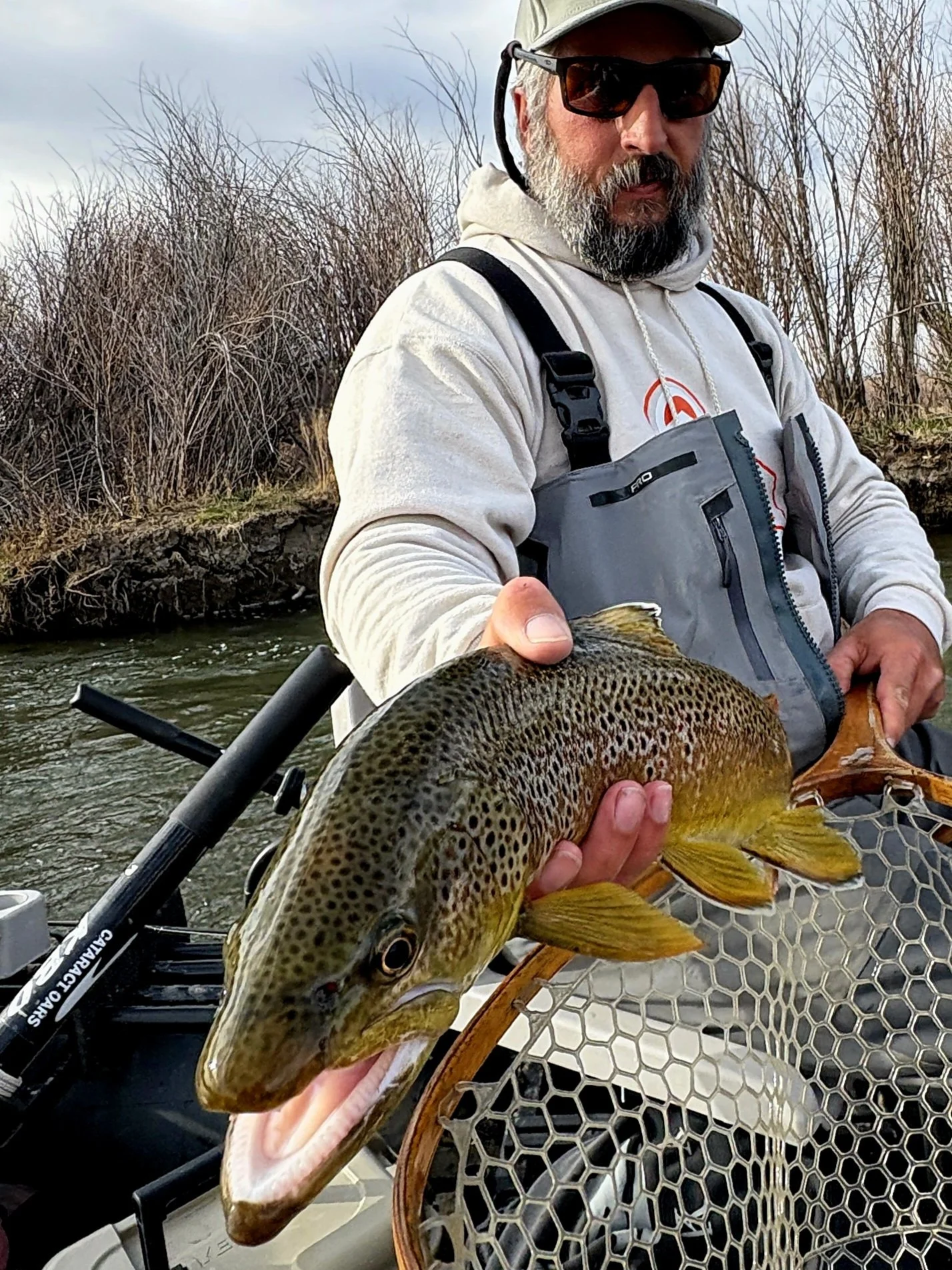 A man with a beard, cap, sunglasses, and outdoor gear holding a large fish over a net, on a boat in a river with leafless bushes in the background.