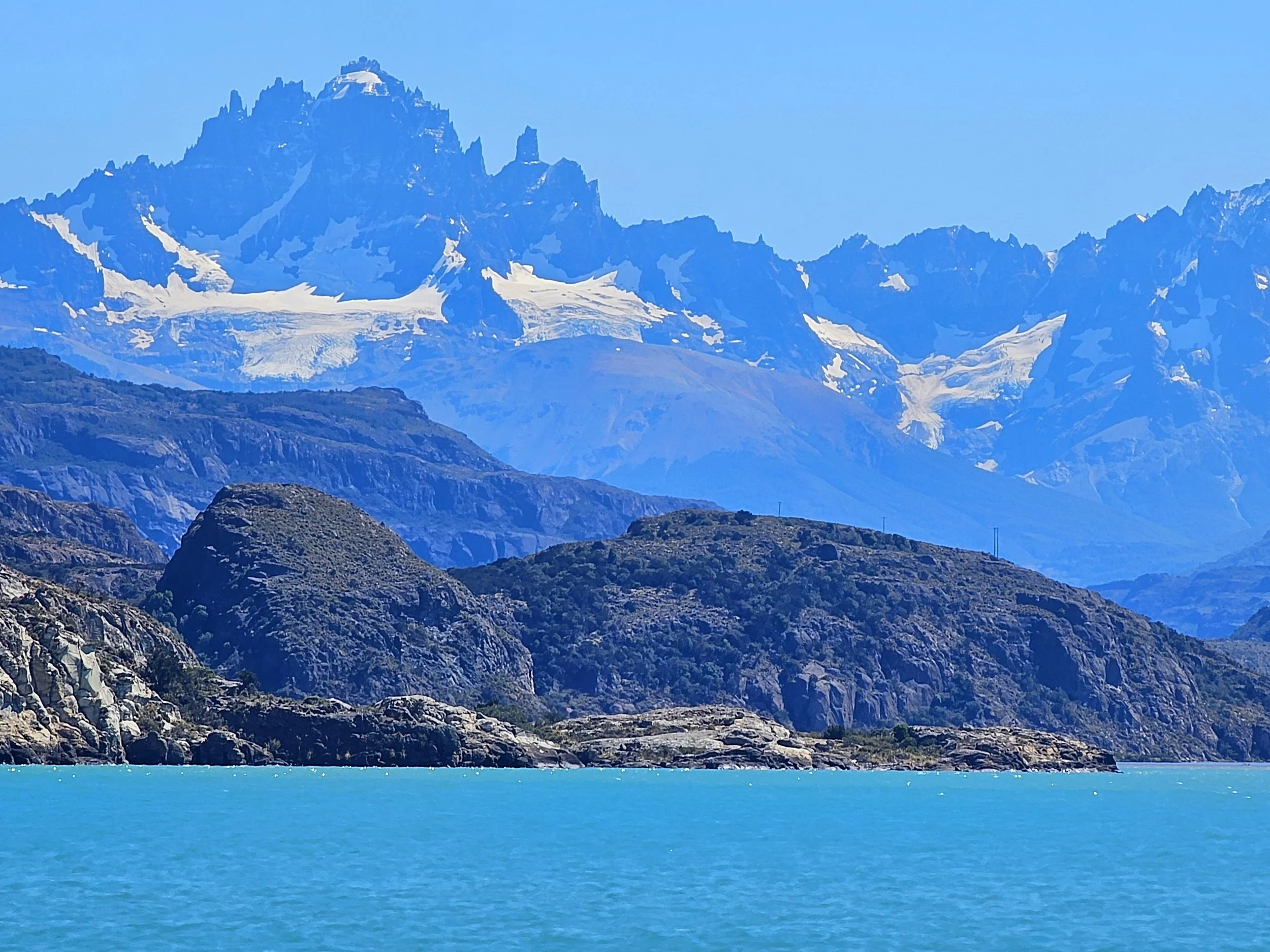 Scenic view of a turquoise lake surrounded by rugged rocky hills, with snow-capped mountains in the background under a clear blue sky.