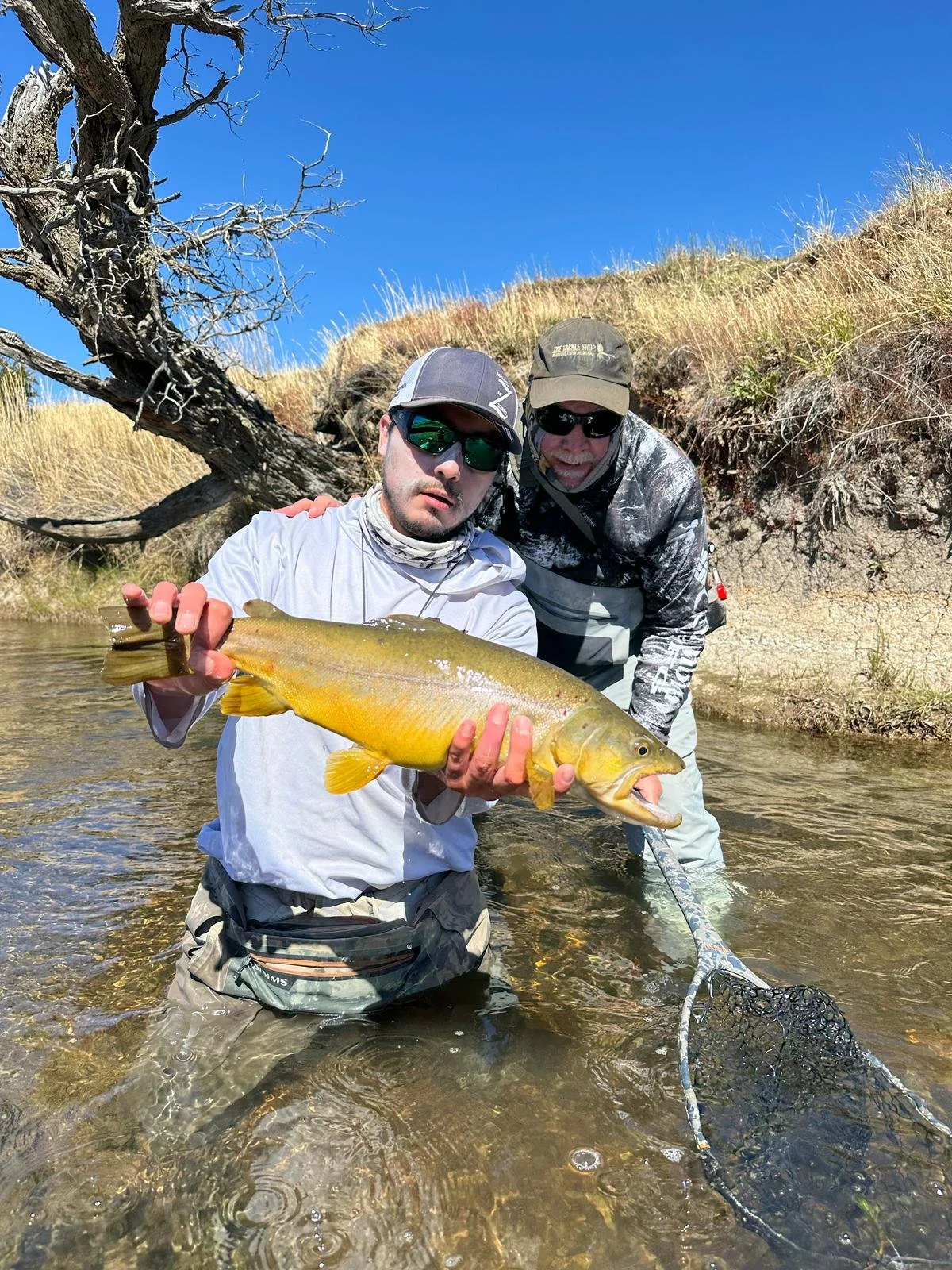 Two men fishing in a river; one showing a large yellow fish with a wide mouth and fins.