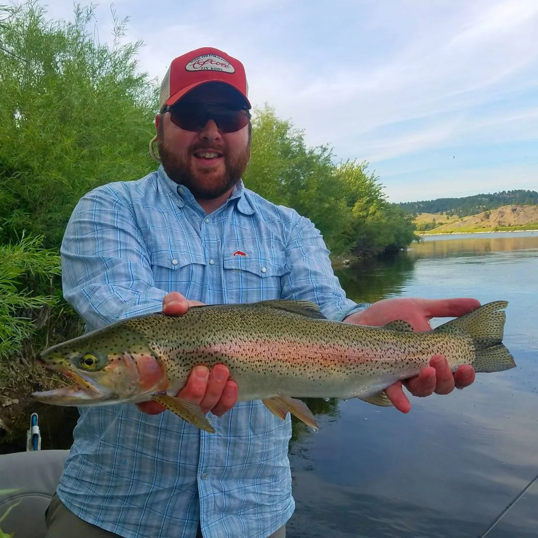 Man wearing sunglasses and a red cap holding a large rainbow trout fish by the river.