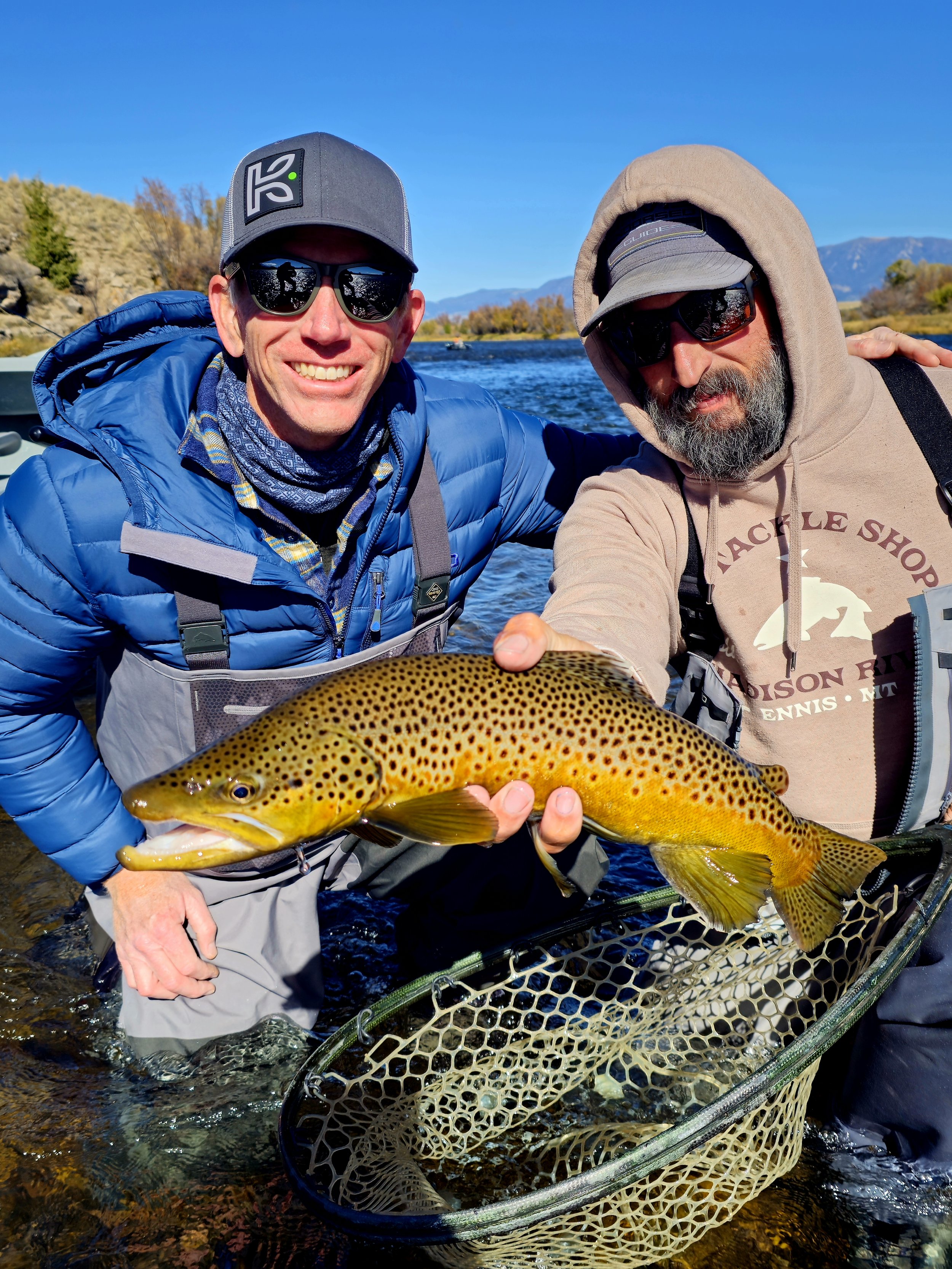 Two men in fishing gear holding a large, spotted fish in a river, with mountains and trees in the background on a sunny day.