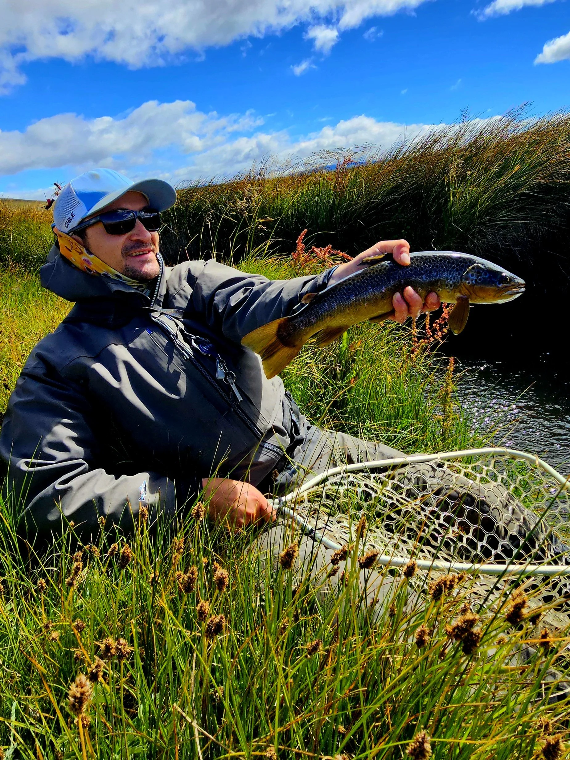 Man holding a large fish near a grassy riverbank under a partly cloudy sky.
