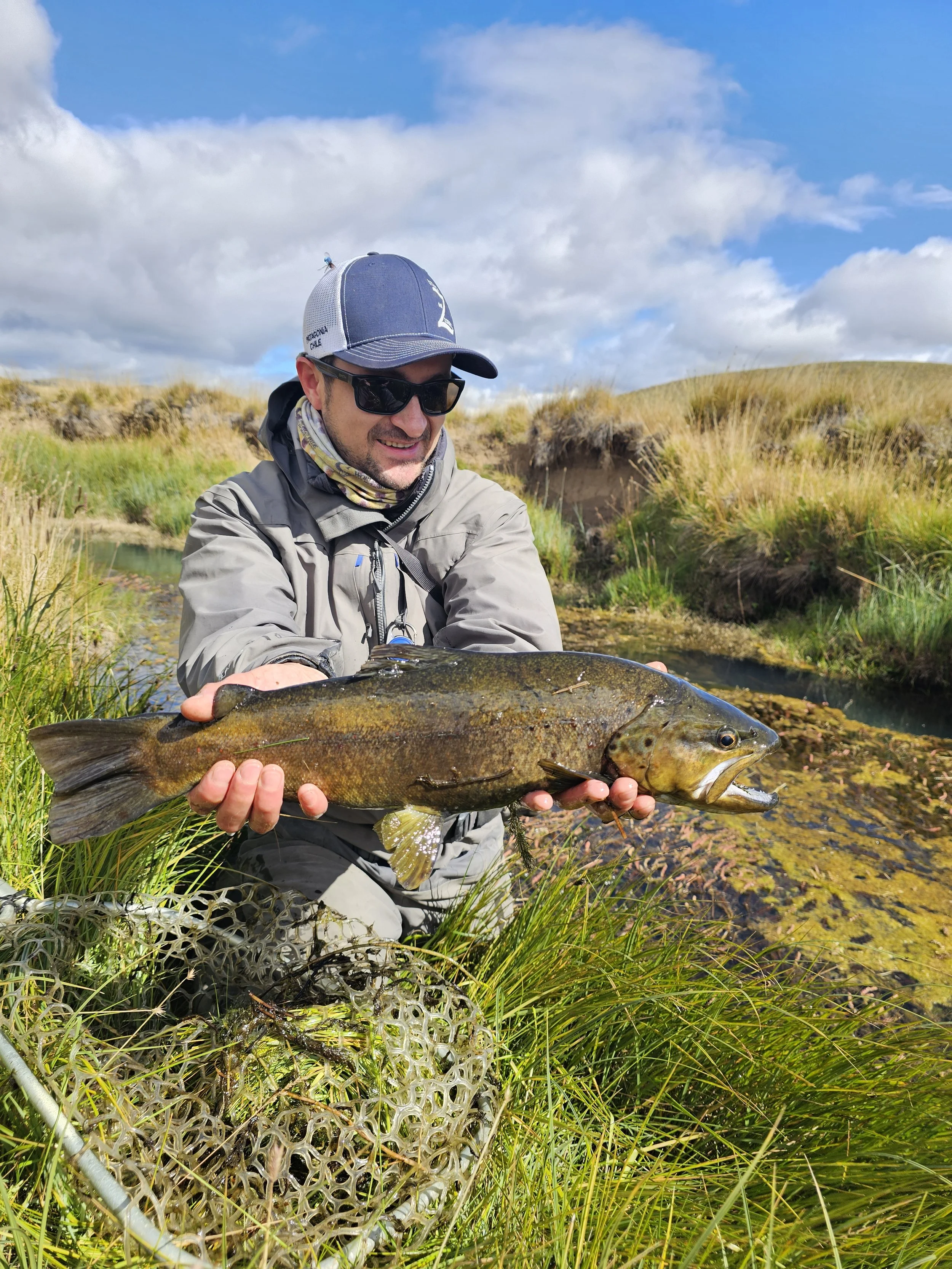 Man wearing sunglasses, cap, and outdoor gear holding a large fish in a grassy and marshy area with blue sky and clouds in the background.