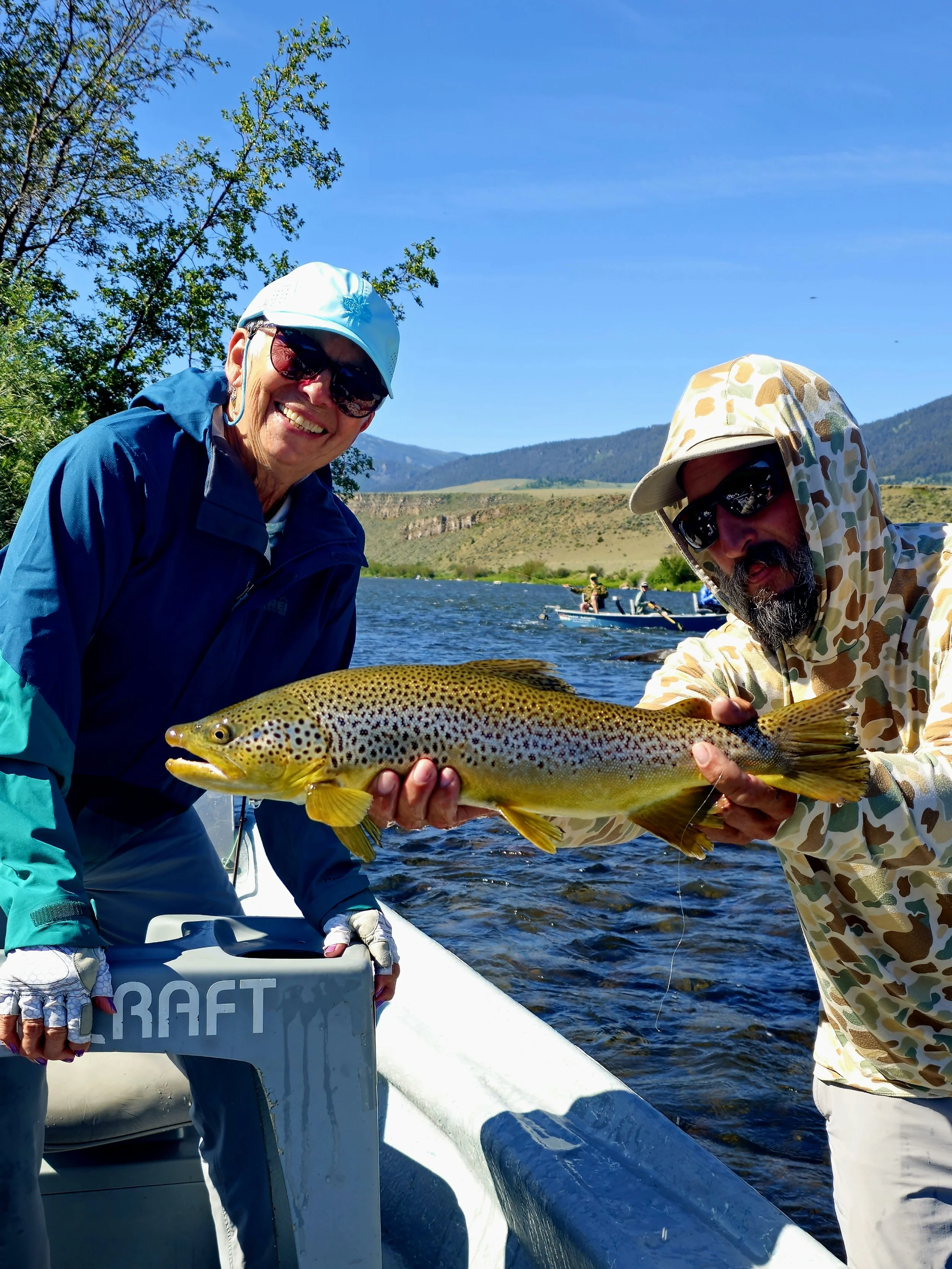 Two people holding a large rainbow trout on a boat, with a waterway and mountains in the background.