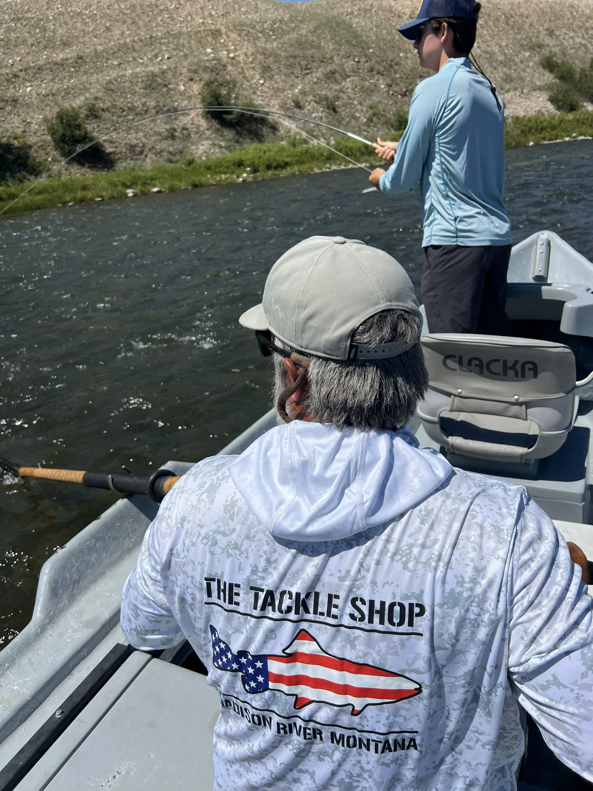 A man and a boy are fishing from a boat on a river with a hillside in the background. The man is wearing a white hoodie with branding from The Tackle Shop in Montana, and the boy is wearing a light blue long-sleeve shirt and a dark cap.