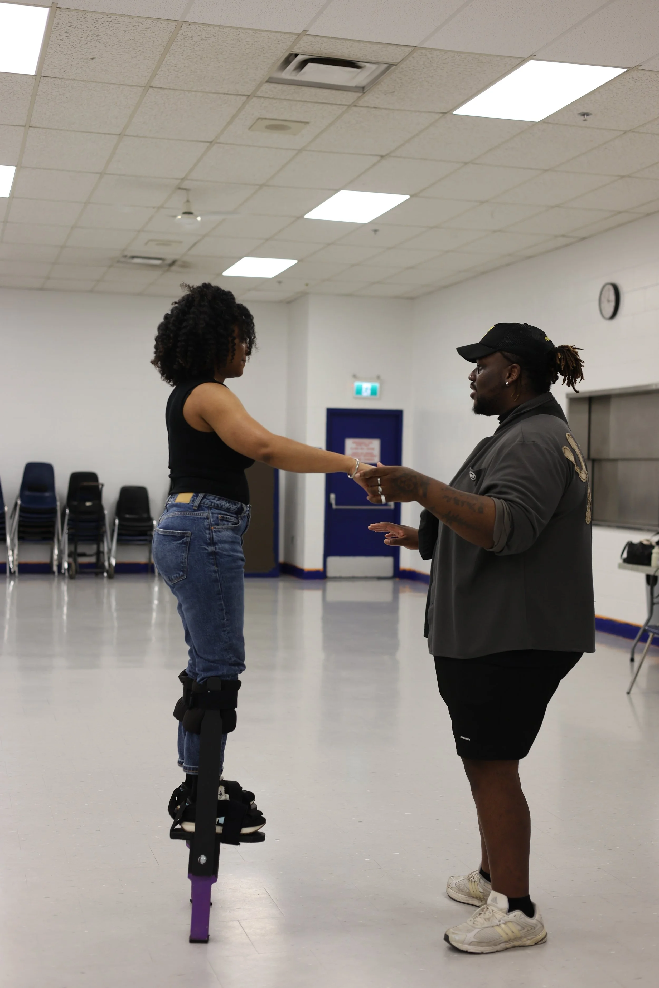 A woman with curly hair on a pair of stilts, holding hands with a man, in a large room with a white ceiling and chairs stacked against the wall.