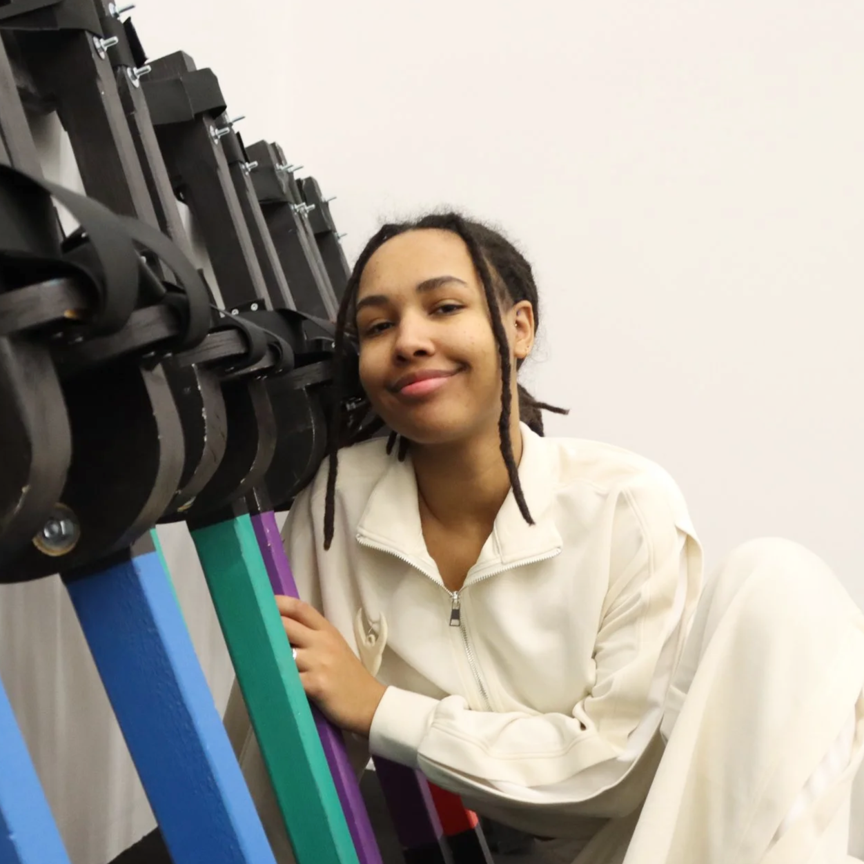 Pixel Heller leaning on a row of stilts, smiling in front of a plain white wall.