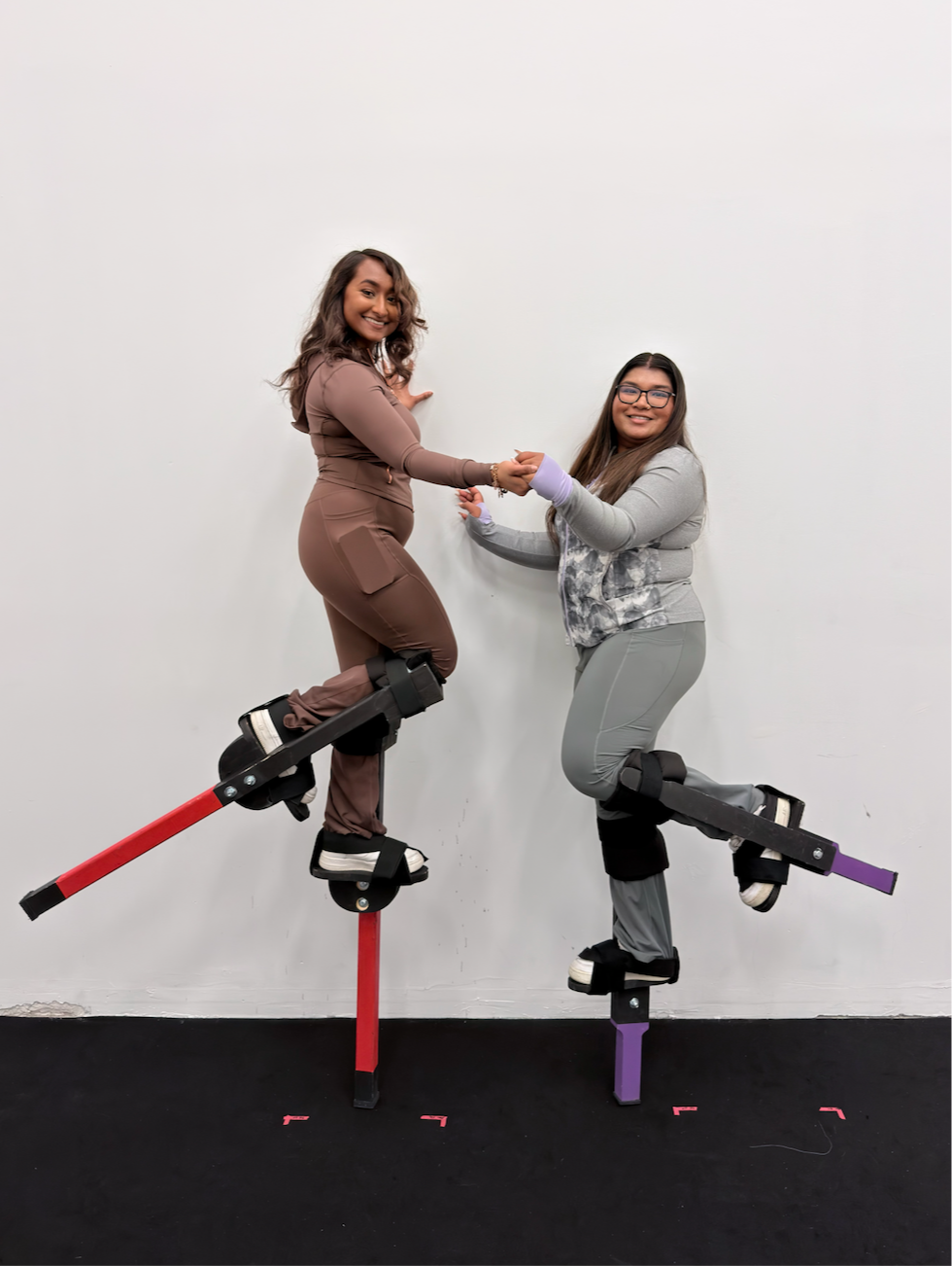 Two women standing on parallel bars and smiling, holding hands, against a plain white wall.