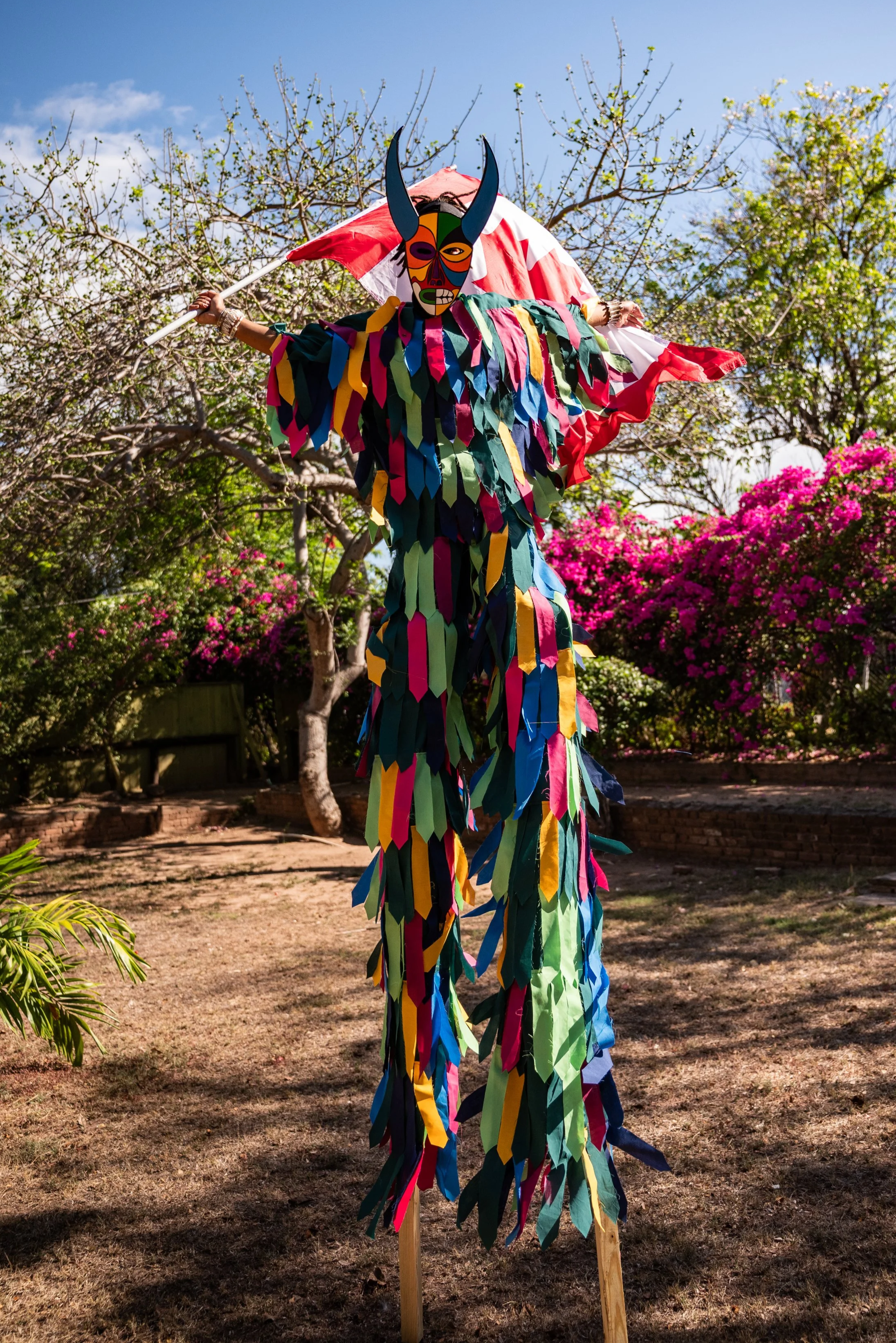 A tall traditional Moko Jumbie with a colorful mask, long colorful costumes, holding the Canadian flag standing outdoors with trees and bushes in the background.