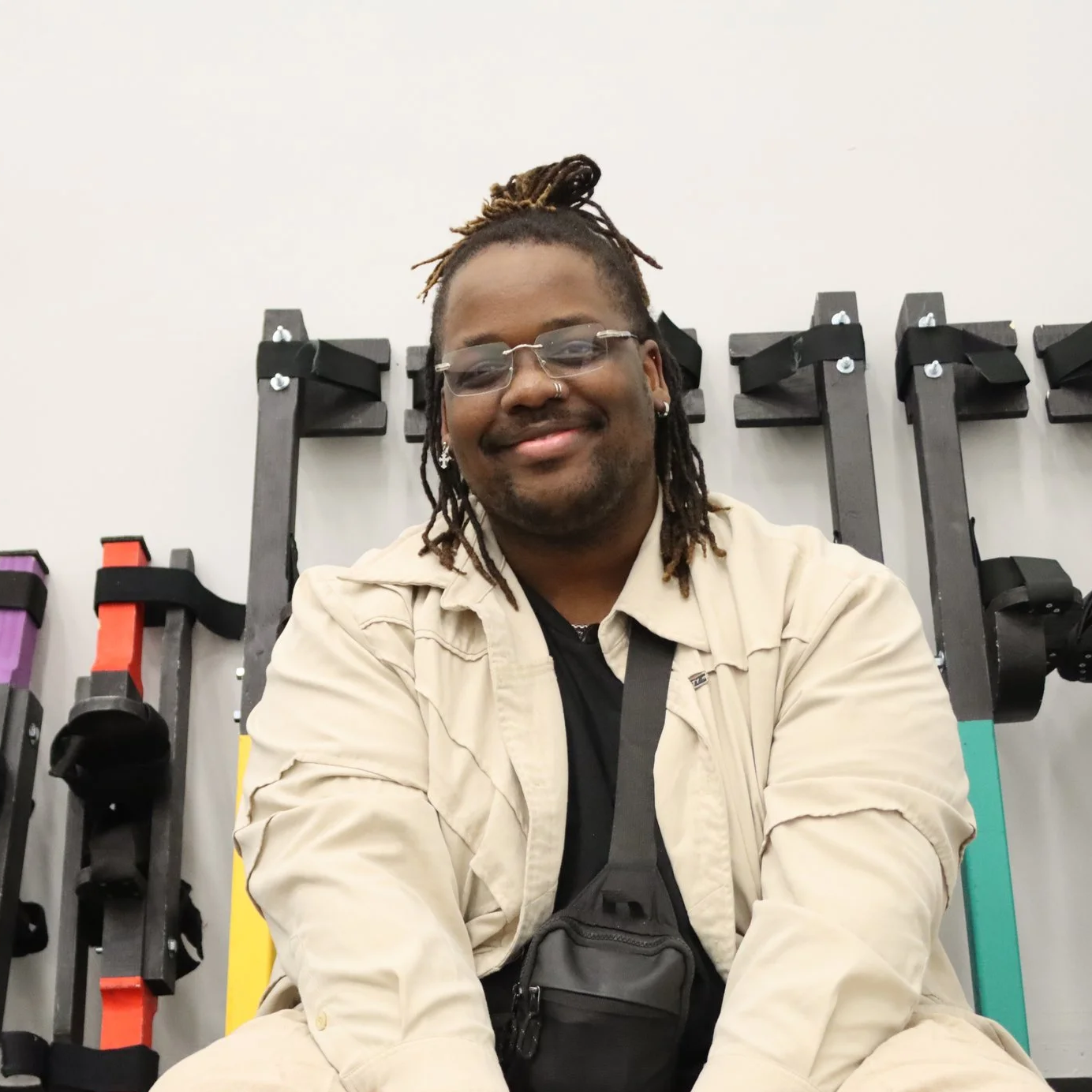 A smiling man with glasses and dreadlocks sitting in front of stilts on the wall.