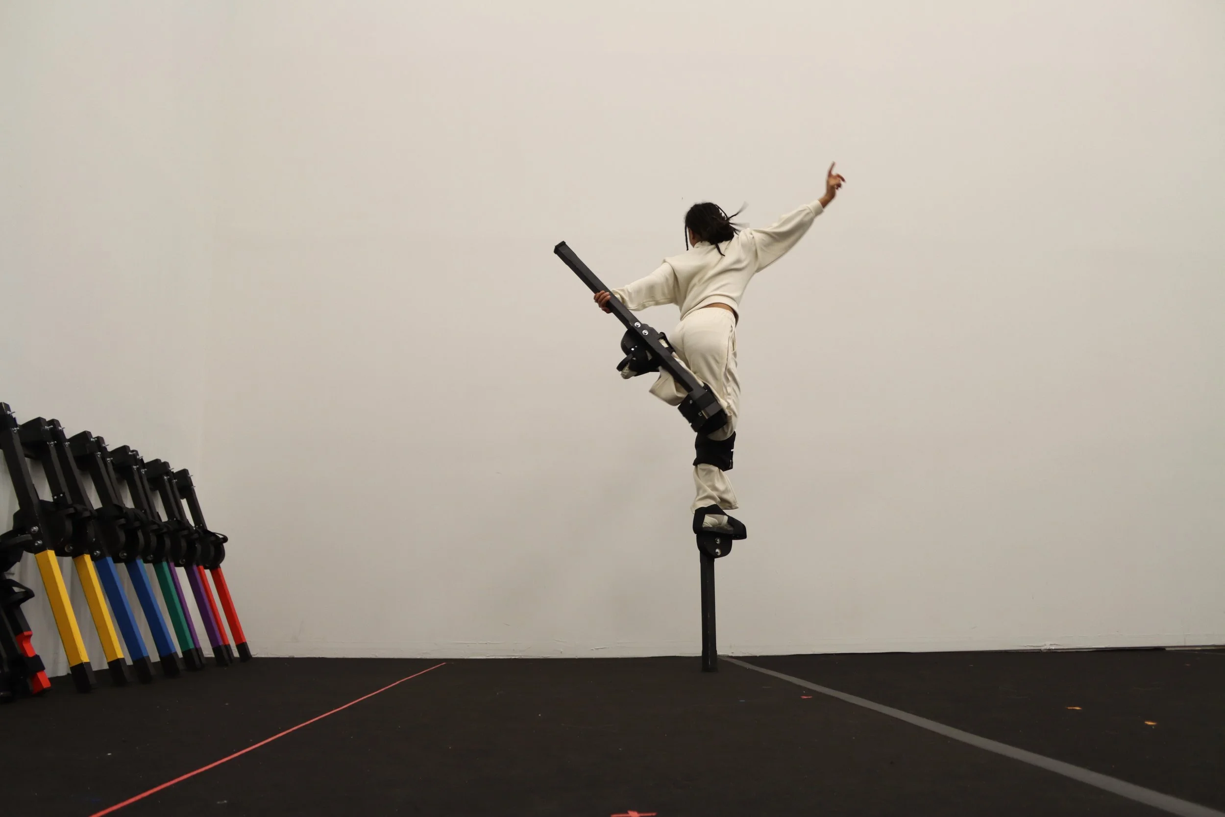 A person balancing on stilts with one leg, holding a large object, in a room with a plain white wall and stacked stilts equipment in the corner.