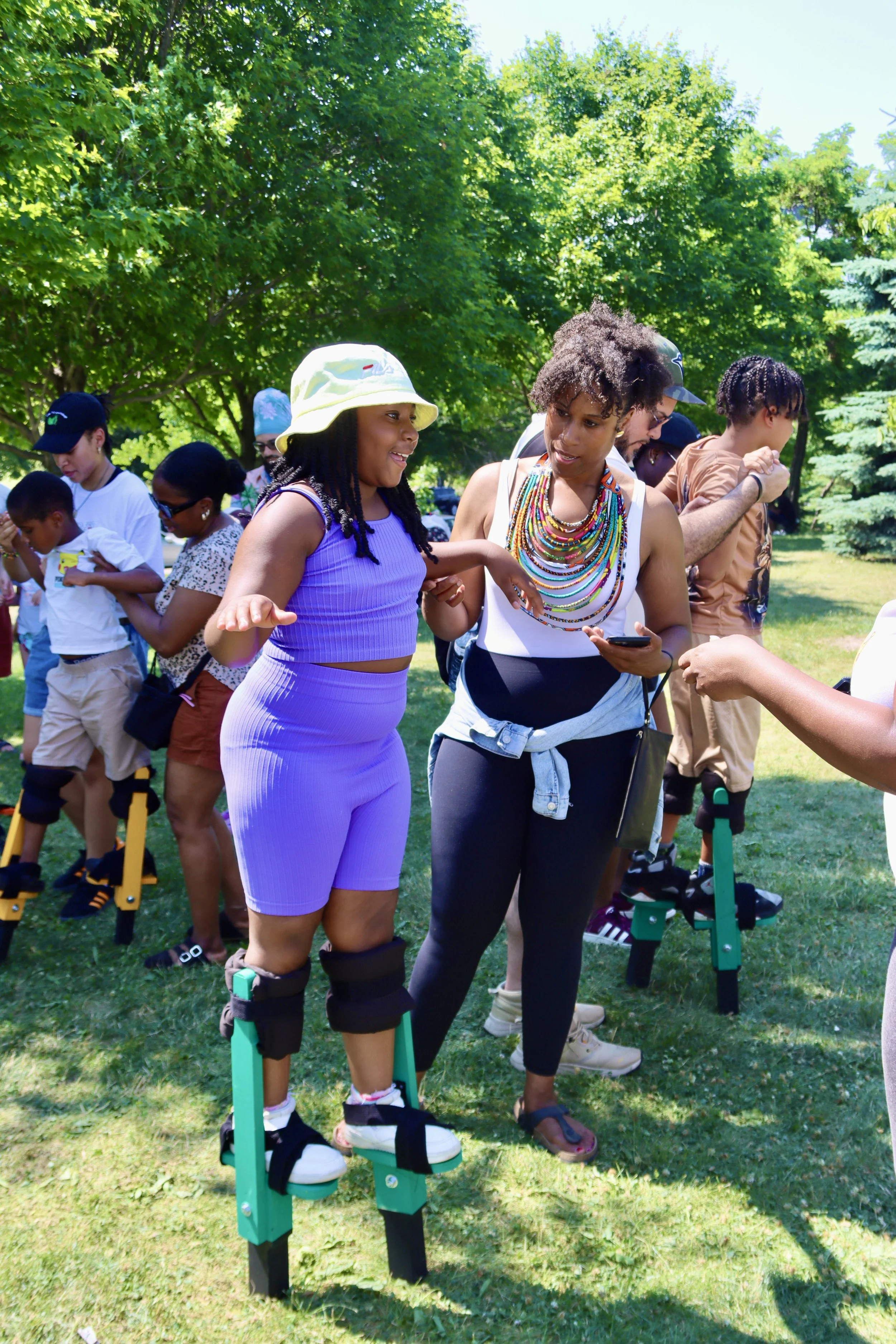 People gathered outside on a sunny day, with a woman and girl in the foreground, the girl wearing stilts and a purple outfit.