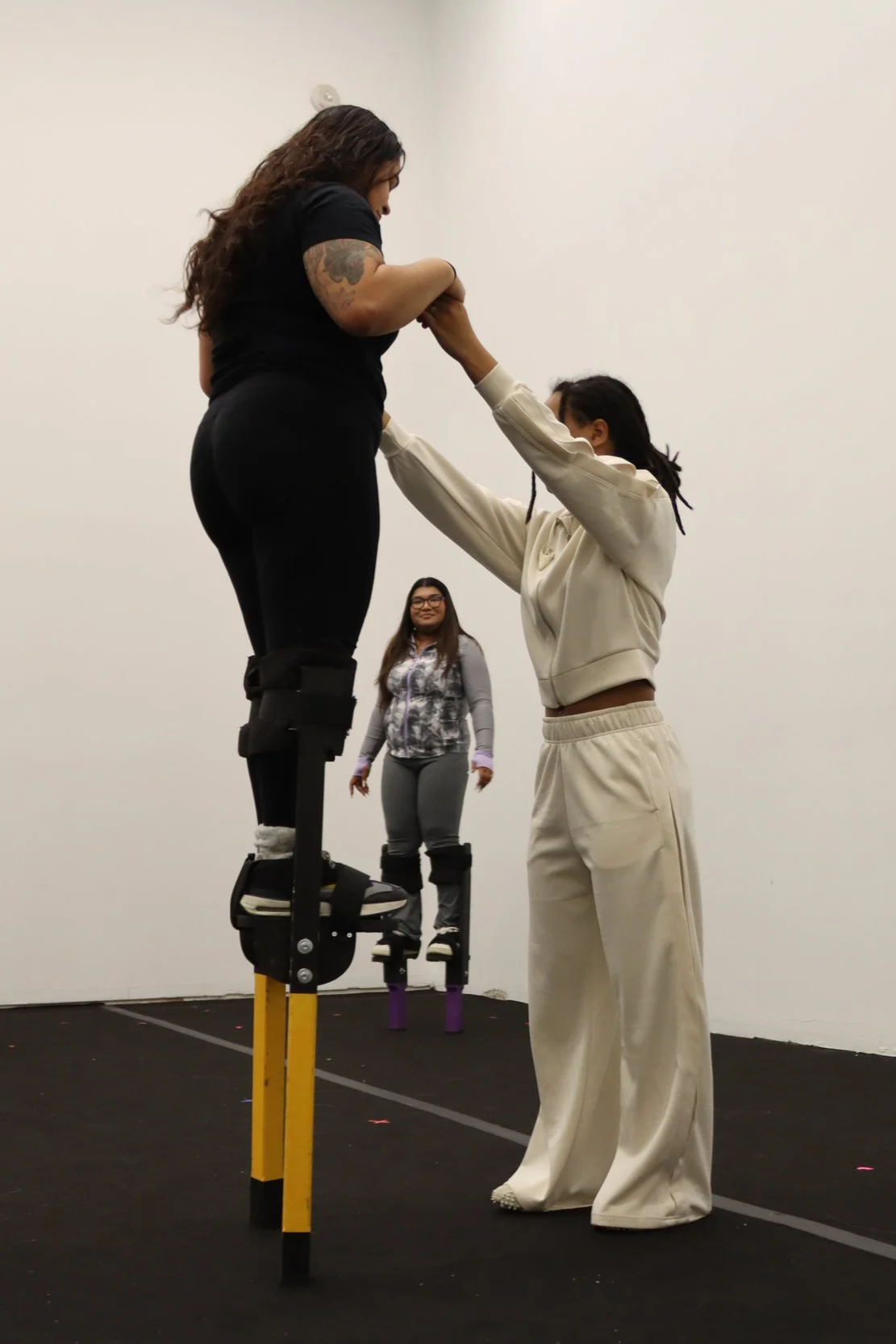 Two women practicing on stilts, one woman holding the other's hands for support while standing on stilts, with a third woman in the background watching and smiling in a plain indoor setting.