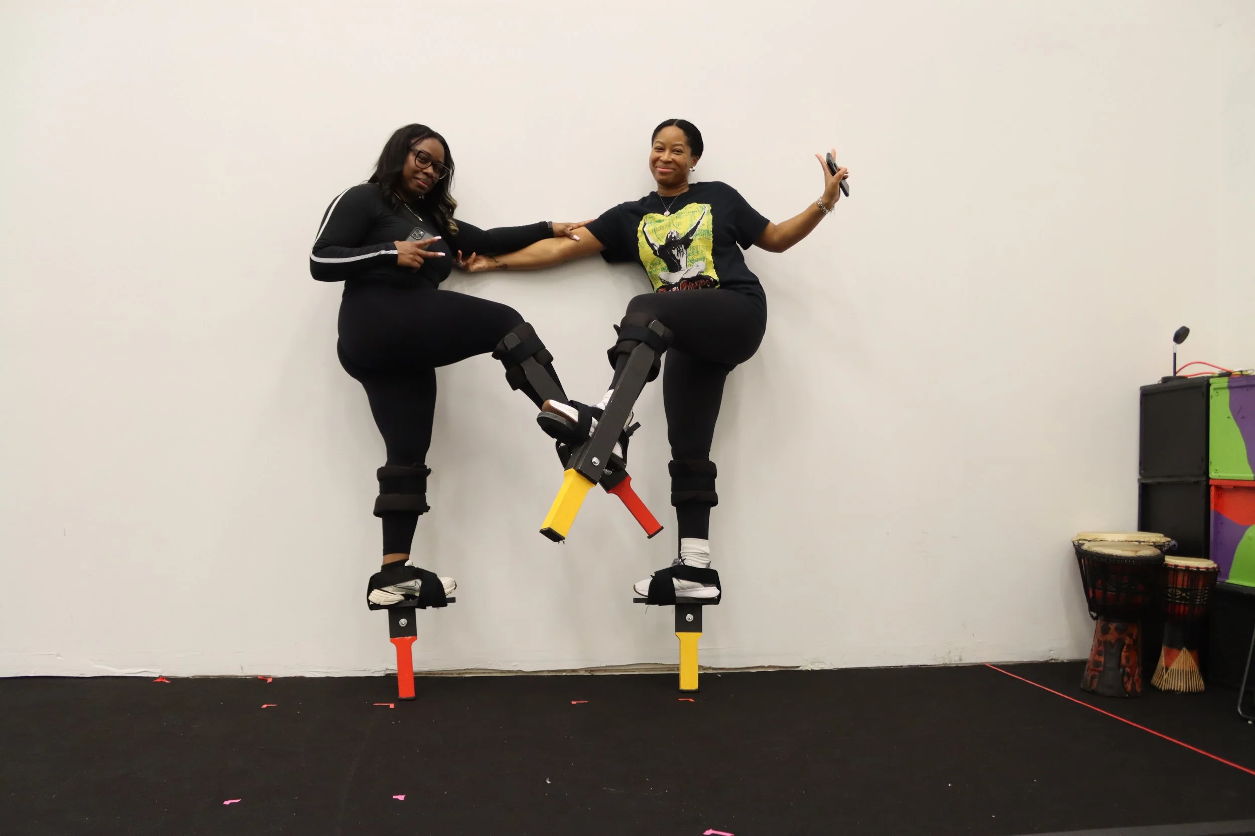 Two women standing on stilts, holding hands for balance, in a room with a plain white wall and music instruments on the side.