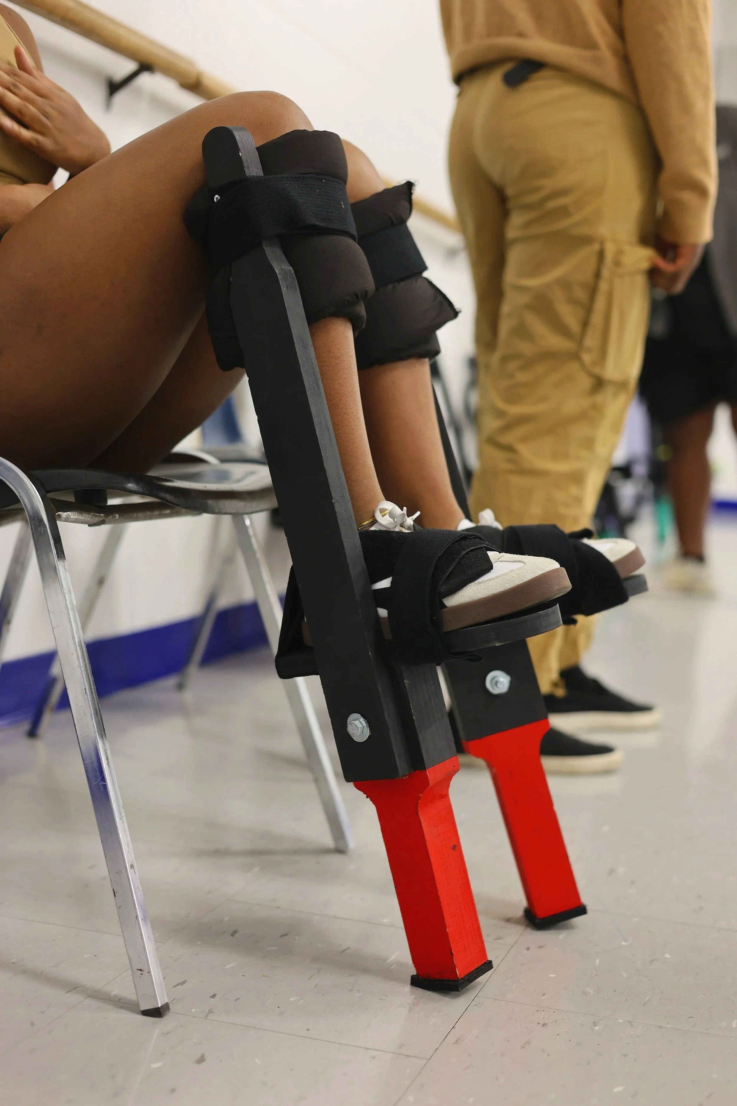 A person is sitting on a chair wearing Moko Jumbie stilts in Ottawa. The person is wearing light-colored sneakers.