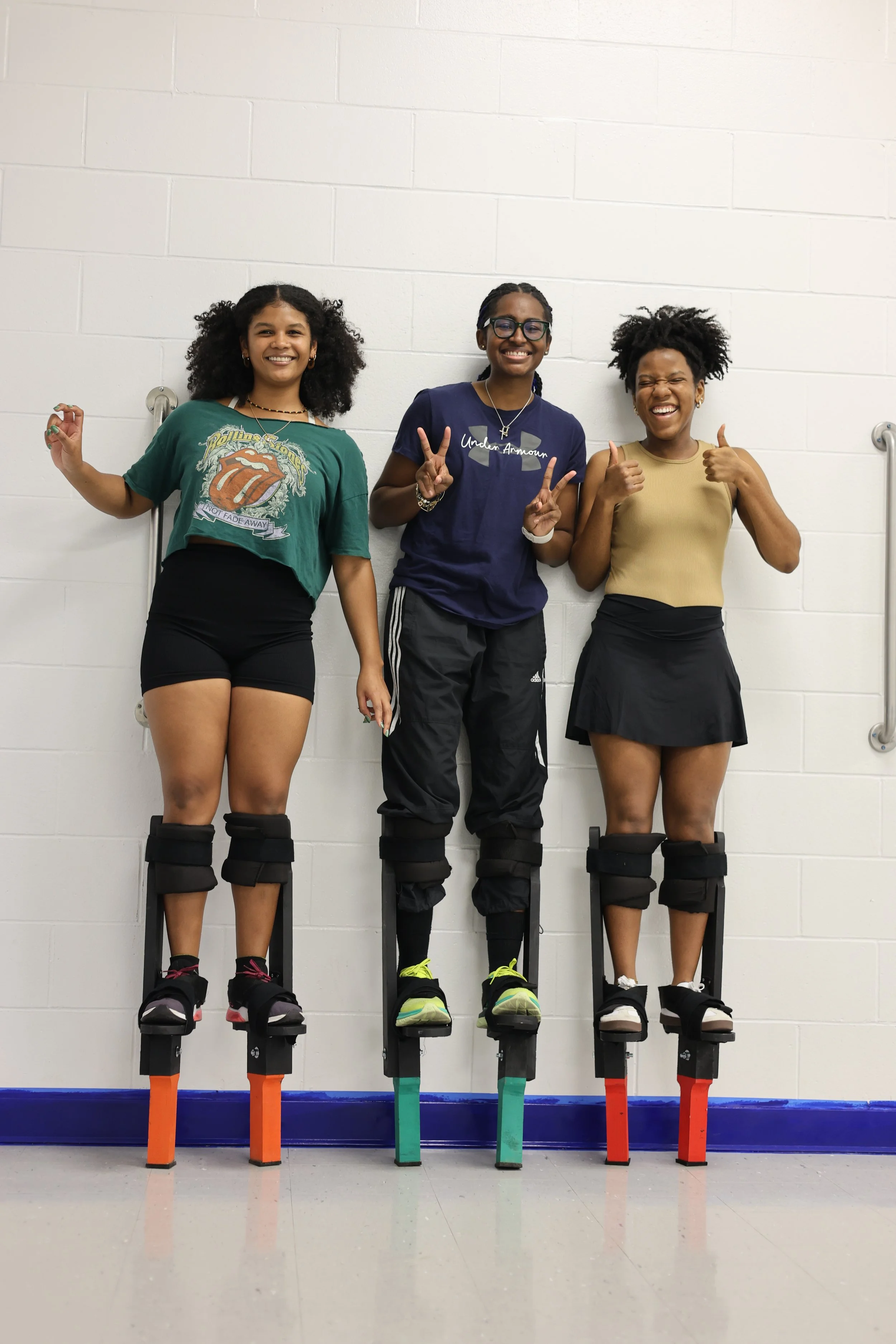 Three women standing on Moko Jumbie stilts, smiling and showing signs of happiness, in a room with a white brick wall.