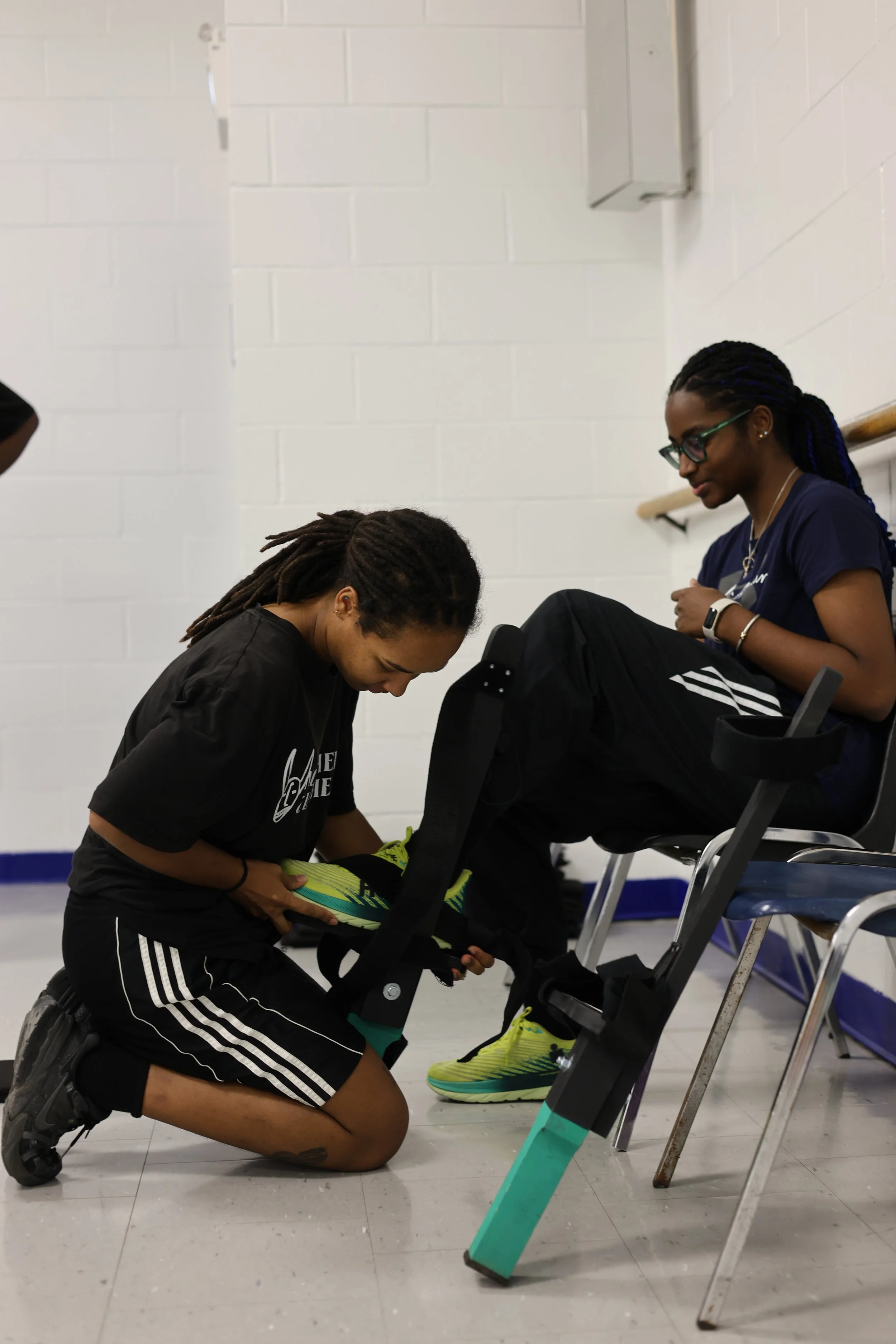 A person kneeling on the floor, adjusting stilts  on another person sitting on a chair against a white cinder block wall.