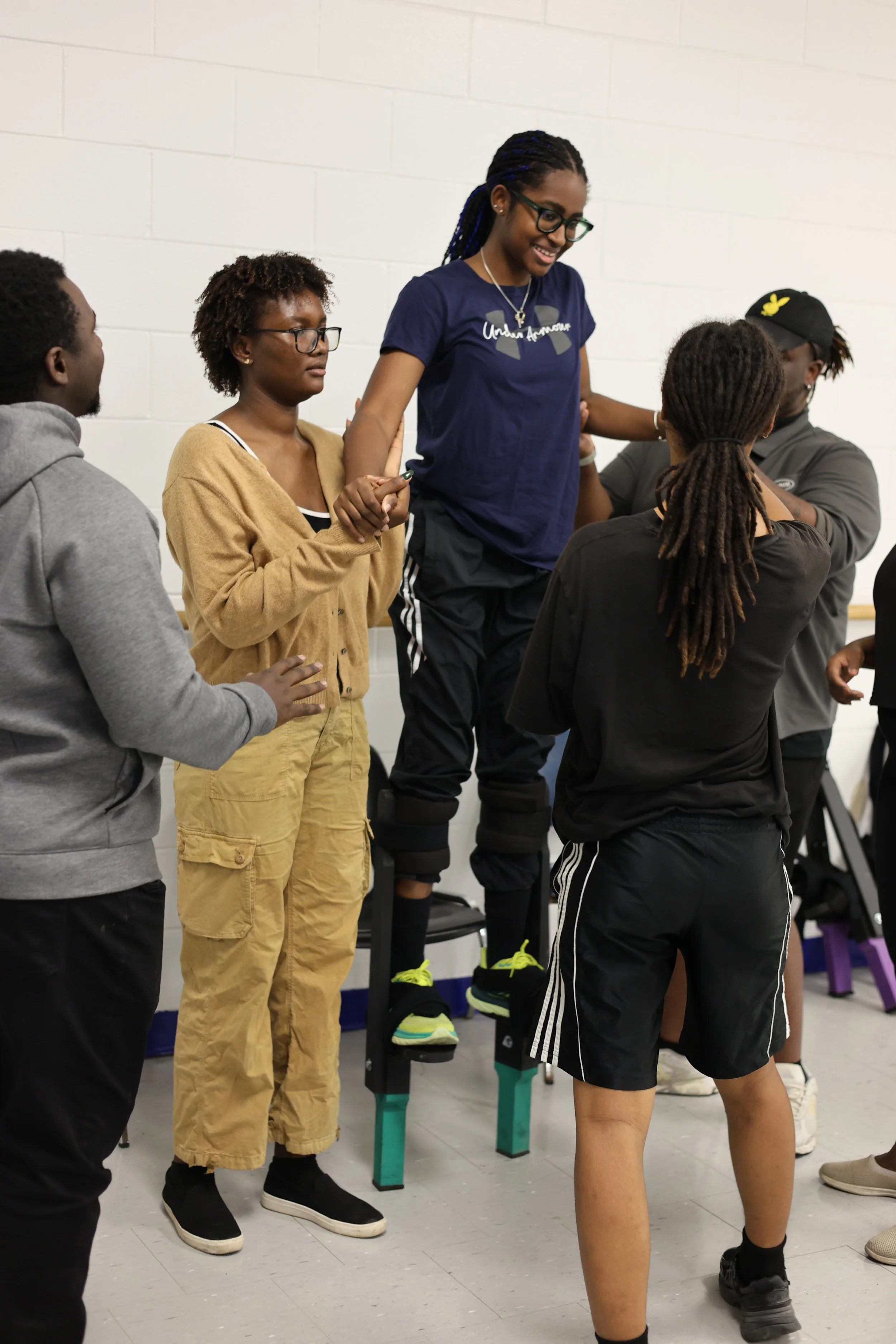 A group of people, including a woman standing on Moko Jumbie stilts, holding hands with others in a circle, in a room with a white brick wall.