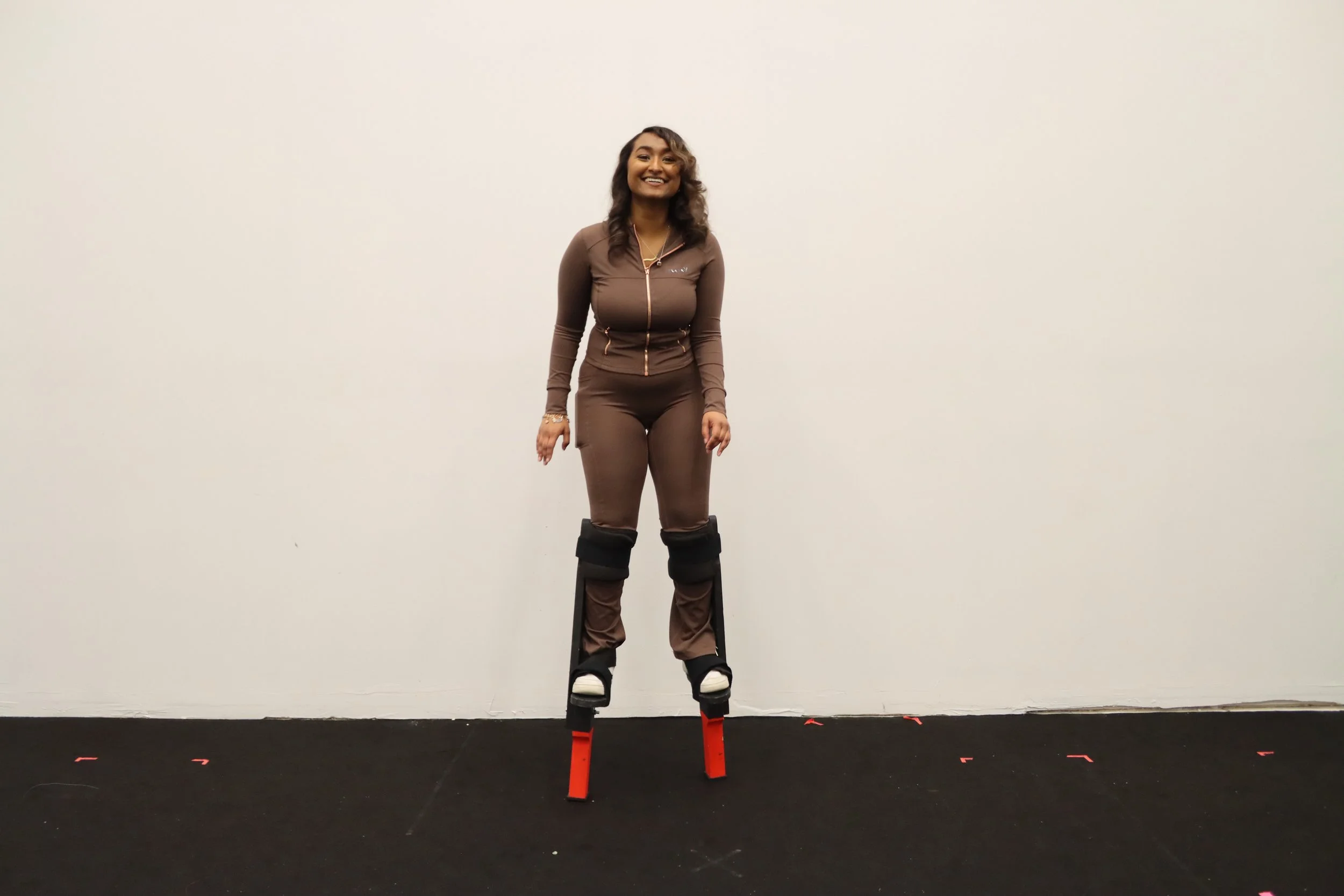 A woman in a brown athletic outfit standing on custom stilts with black straps, in a room with a plain white wall and black flooring.