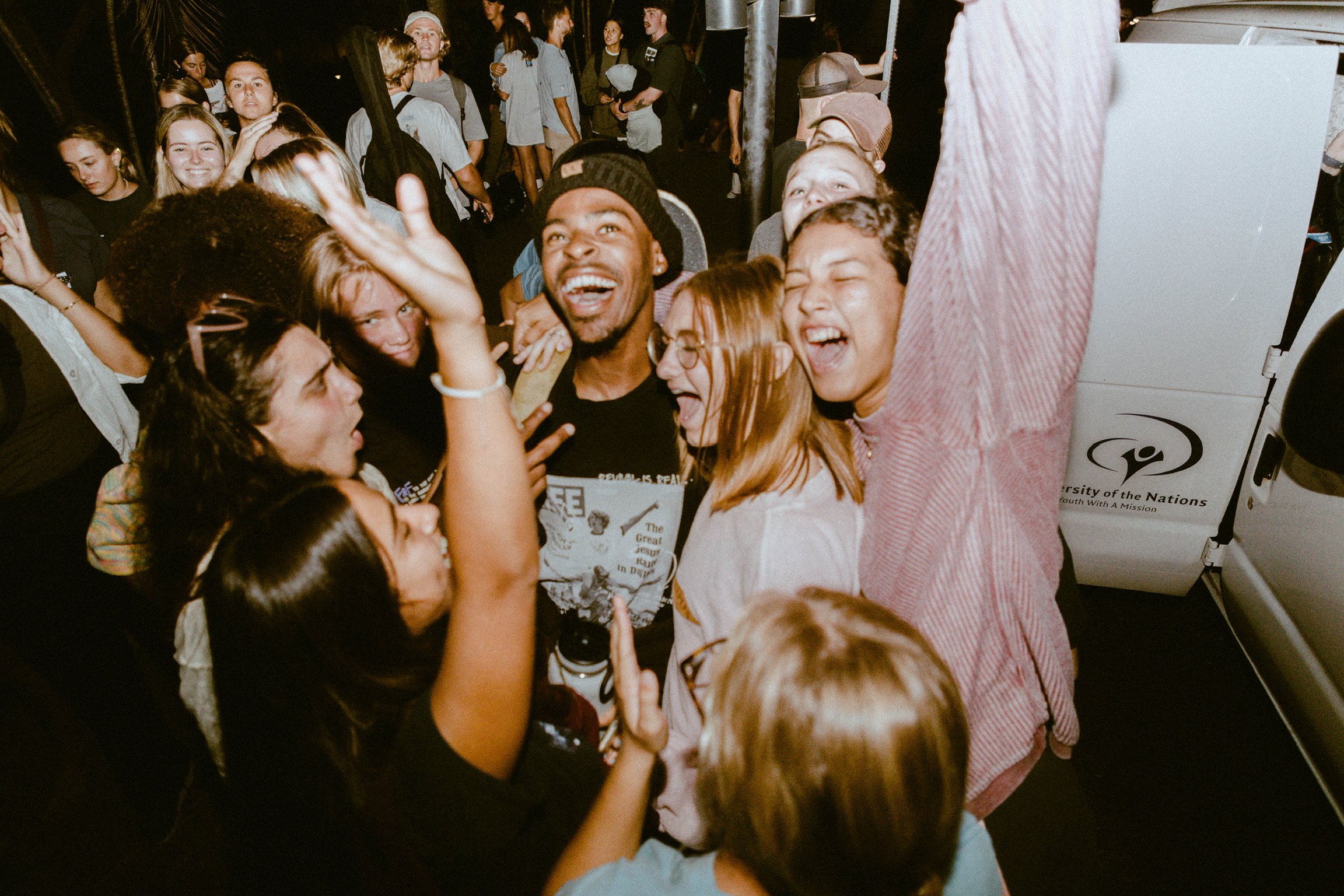 Group of diverse young people smiling, laughing, and taking selfies at a lively event, with some in the background.