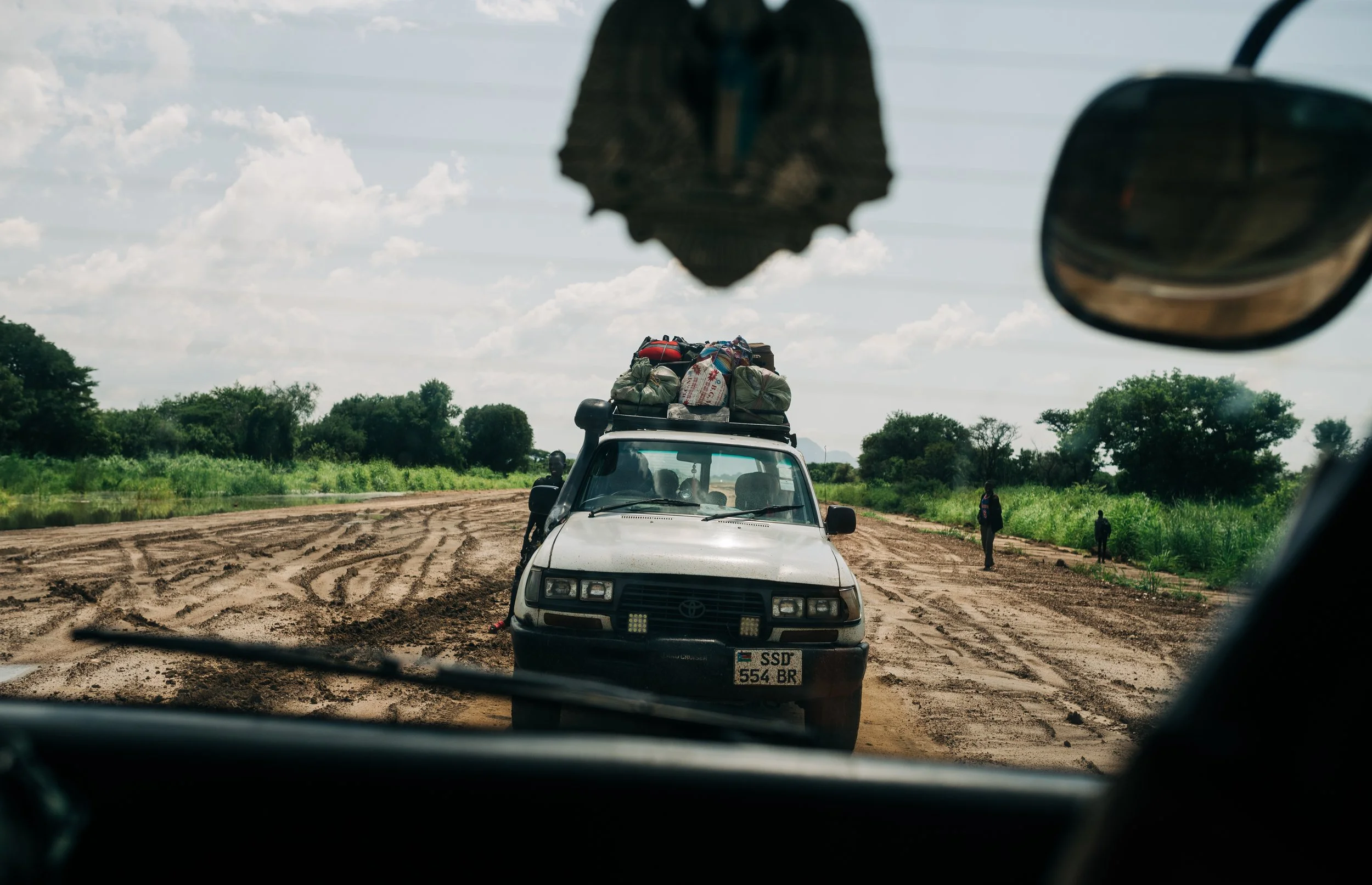 View from inside a vehicle showing a muddy road ahead with a white pickup truck loaded with luggage and supplies on top, surrounded by green trees and a partly cloudy sky, along with people walking on the side of the road.