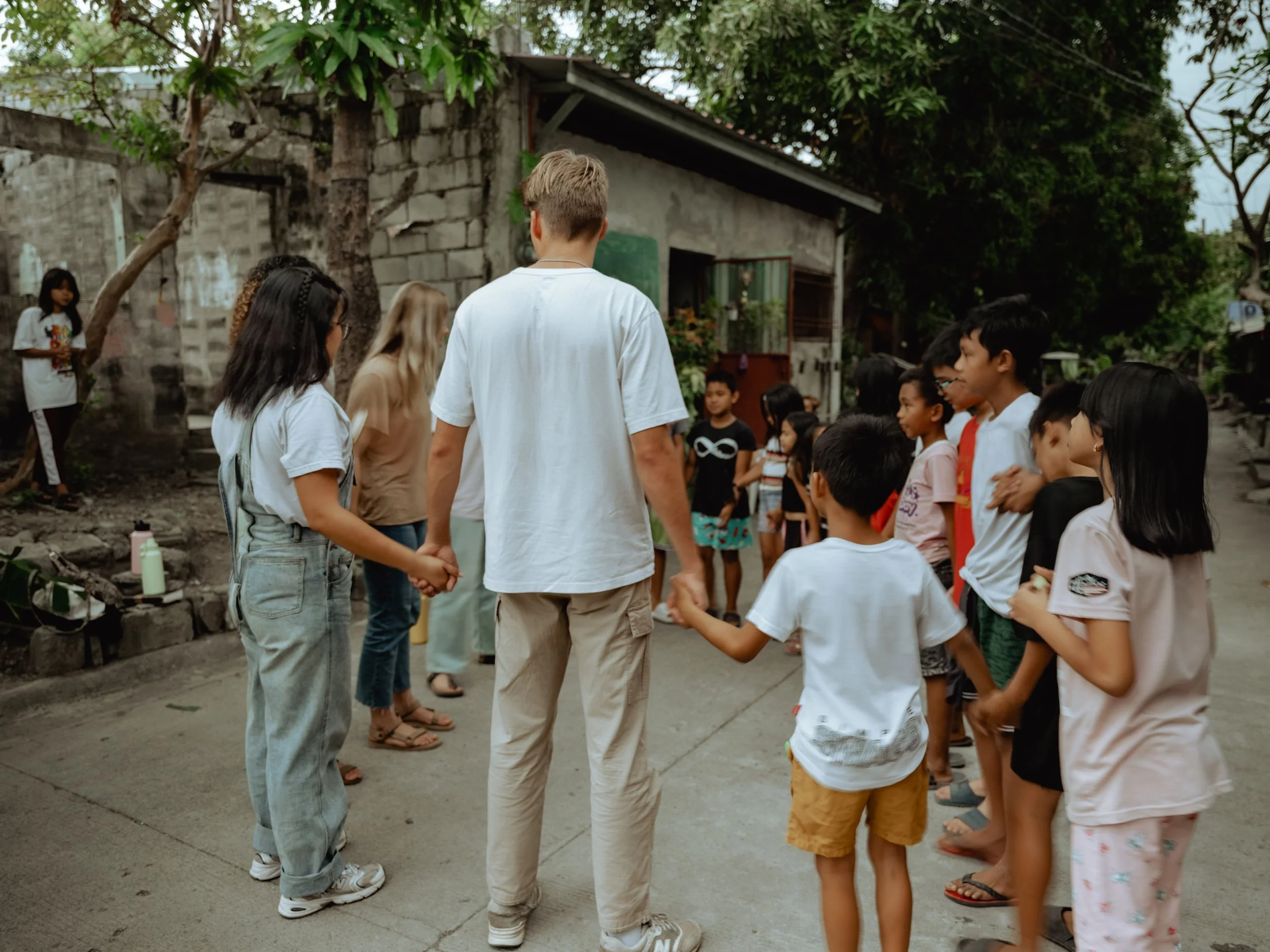 A diverse group of children and teenagers are holding hands in a circle outdoors, participating in a gathering or activity, with a rustic background of trees and an old building.