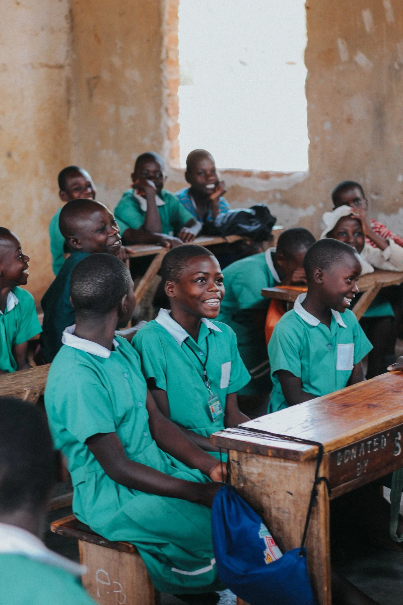 Children sitting in a classroom, wearing green uniforms, smiling and paying attention.