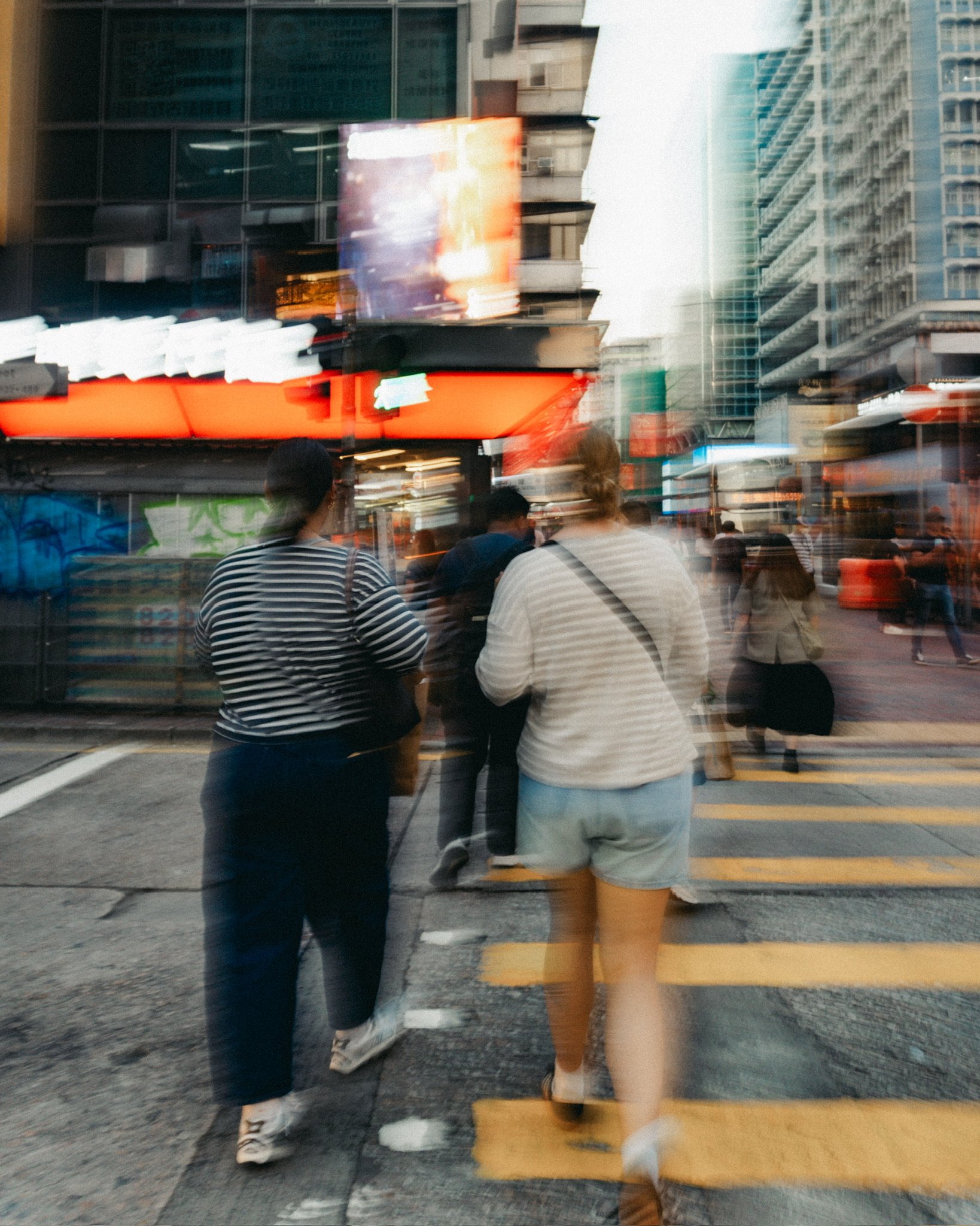 People crossing the street in an urban area with tall buildings and bright neon lights, captured with motion blur.