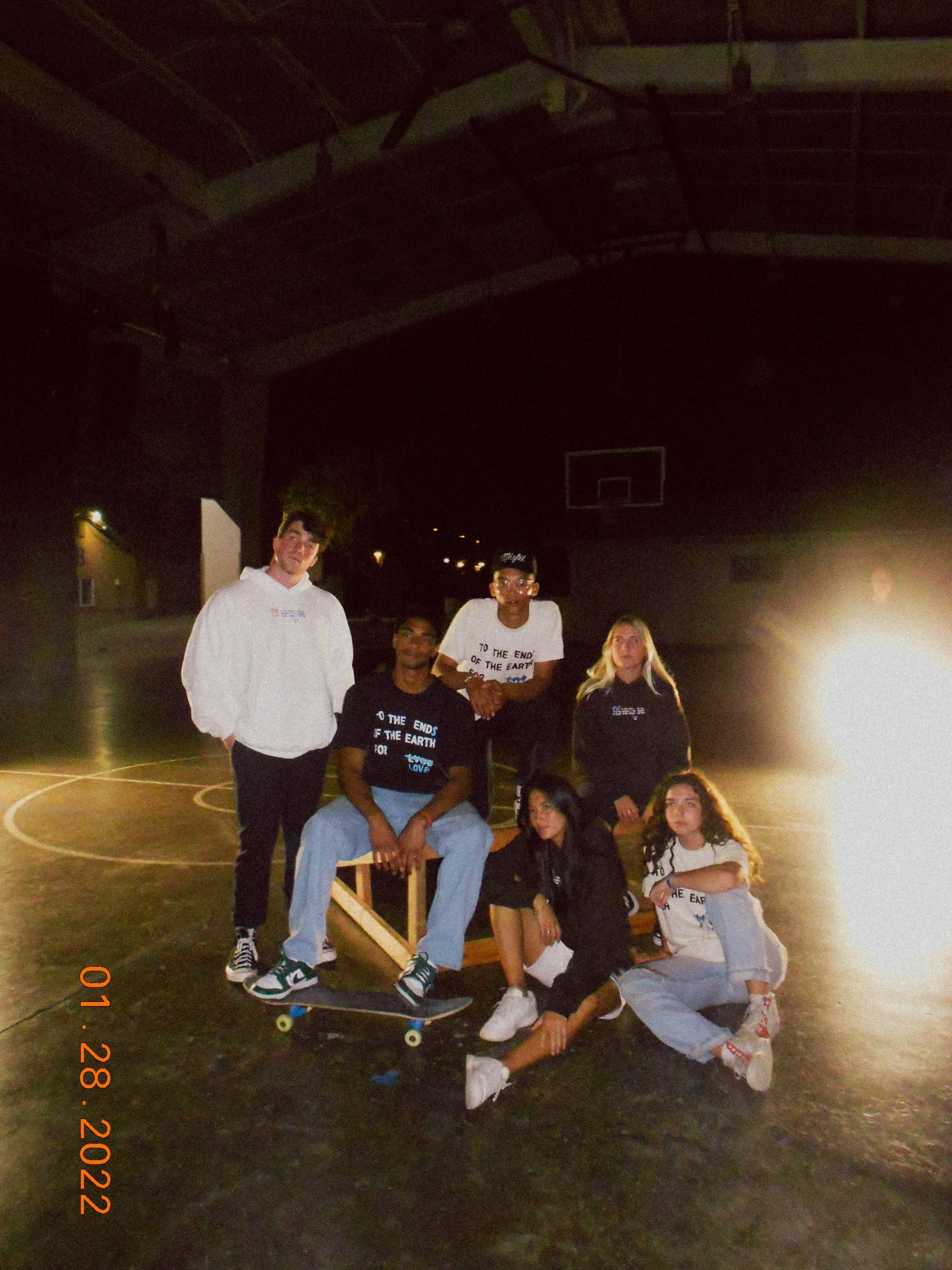 Group of six young people posing on a basketball court at night, with one sitting on a skateboard, under a dark roof, some wearing black T-shirts with the text 'To the End of the Earth'.