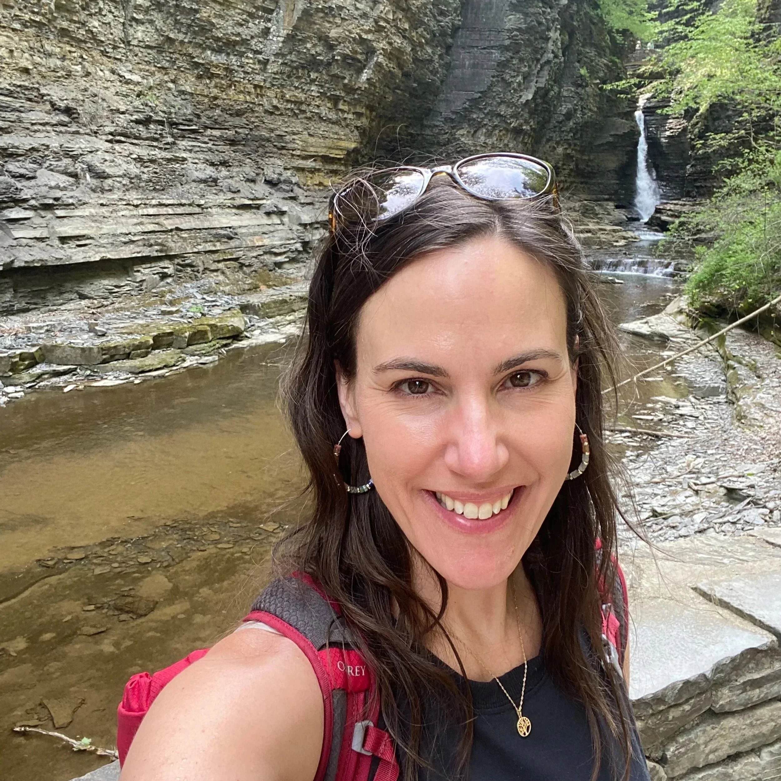 A woman smiling with a waterfall and rocky waterfall in the background, wearing a black shirt, red backpack, and sunglasses on her head. Baltimore chiropractor, gentle chiropractic, kids and pregnancy, family chiropractic, chiropractor near me