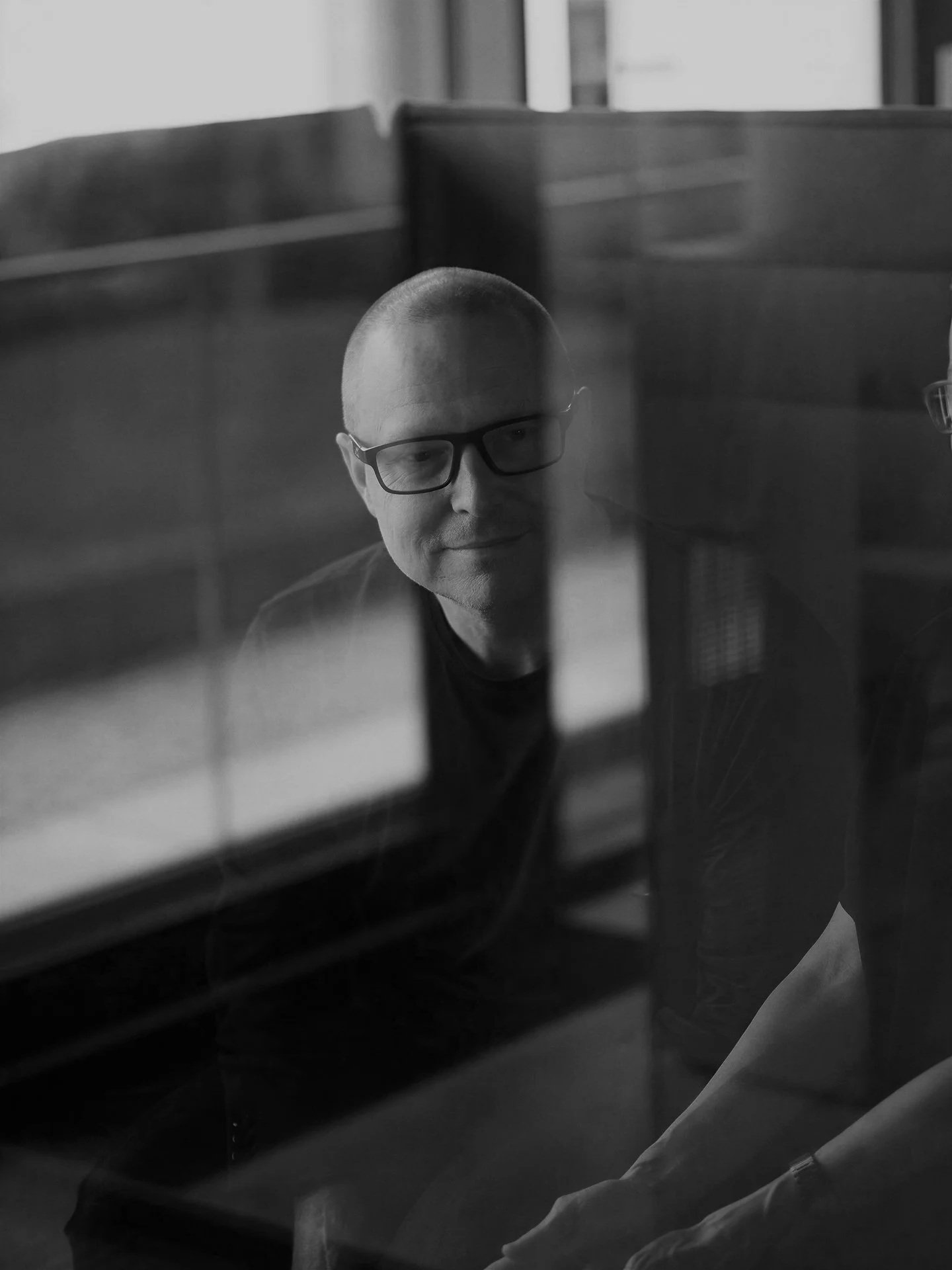 A man with glasses and a shaved head viewed through a glass window, with reflections of a room and objects inside.