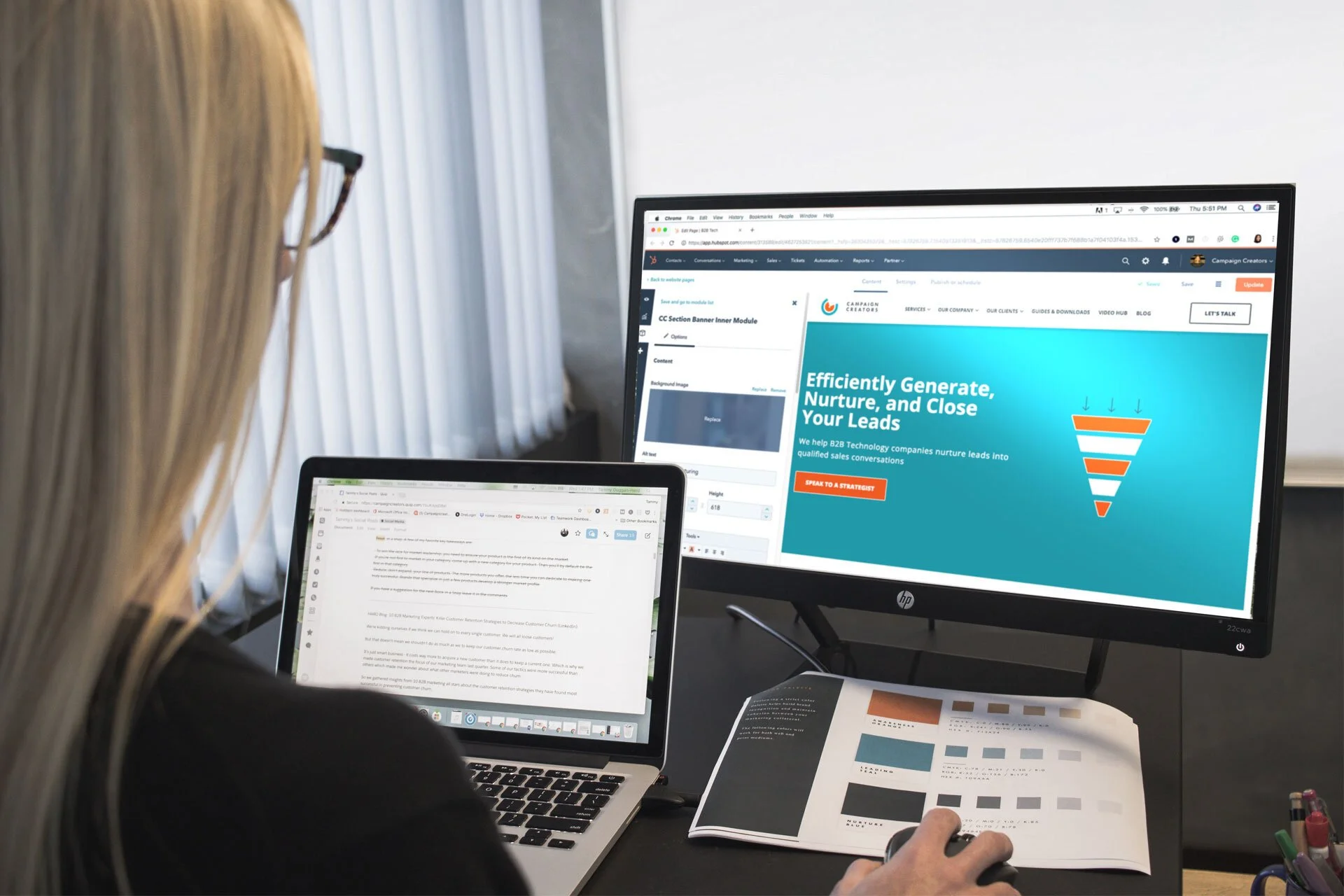 A person with long blonde hair and glasses works at a desk with a laptop and a large monitor. The laptop screen displays text, and the monitor shows a website related to generating and nurturing leads for sales. There is also an open color palette or sample sheet on the desk.