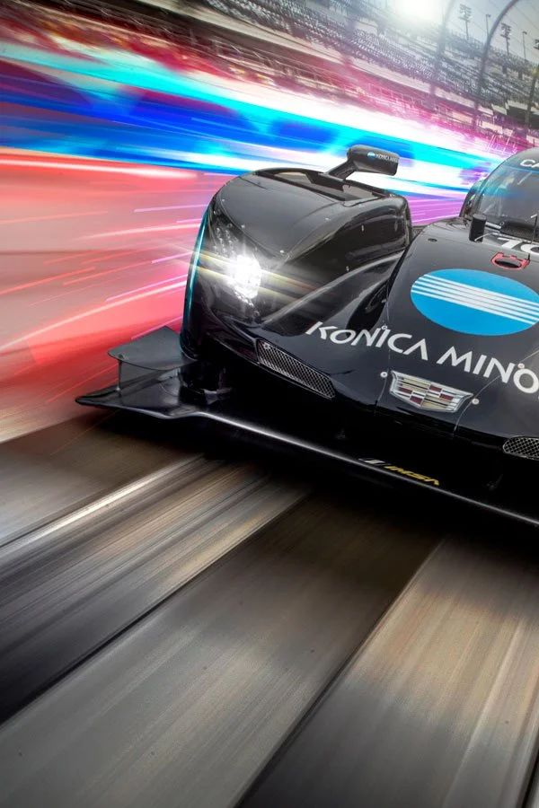 A black racing car with sponsorship logos on a race track, with streaks of colorful lights in the background indicating high speed.