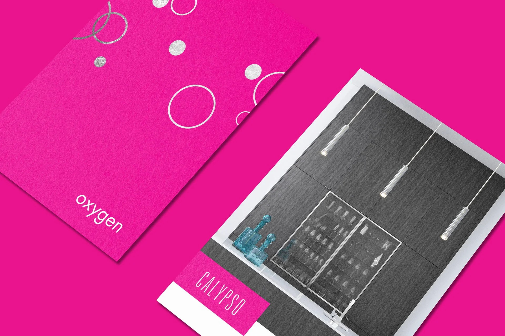 Close-up of two pink cards on a bright pink surface, one with white circular designs and the word "oxygen", the other with a photo of kitchen cabinets and a kitchen scene, labeled "CALIPSO".
