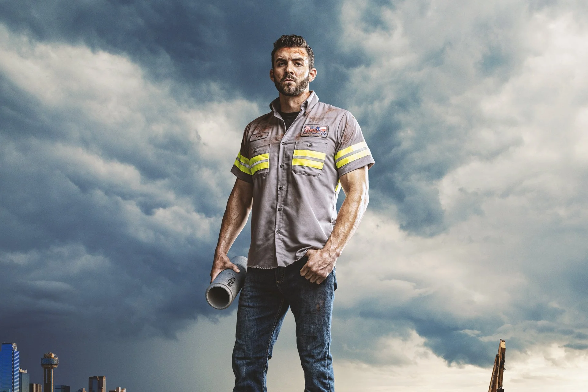 A construction worker wearing a gray uniform with yellow reflective stripes, holding a rolled-up blueprint, standing against a cloudy sky with a city skyline in the background.