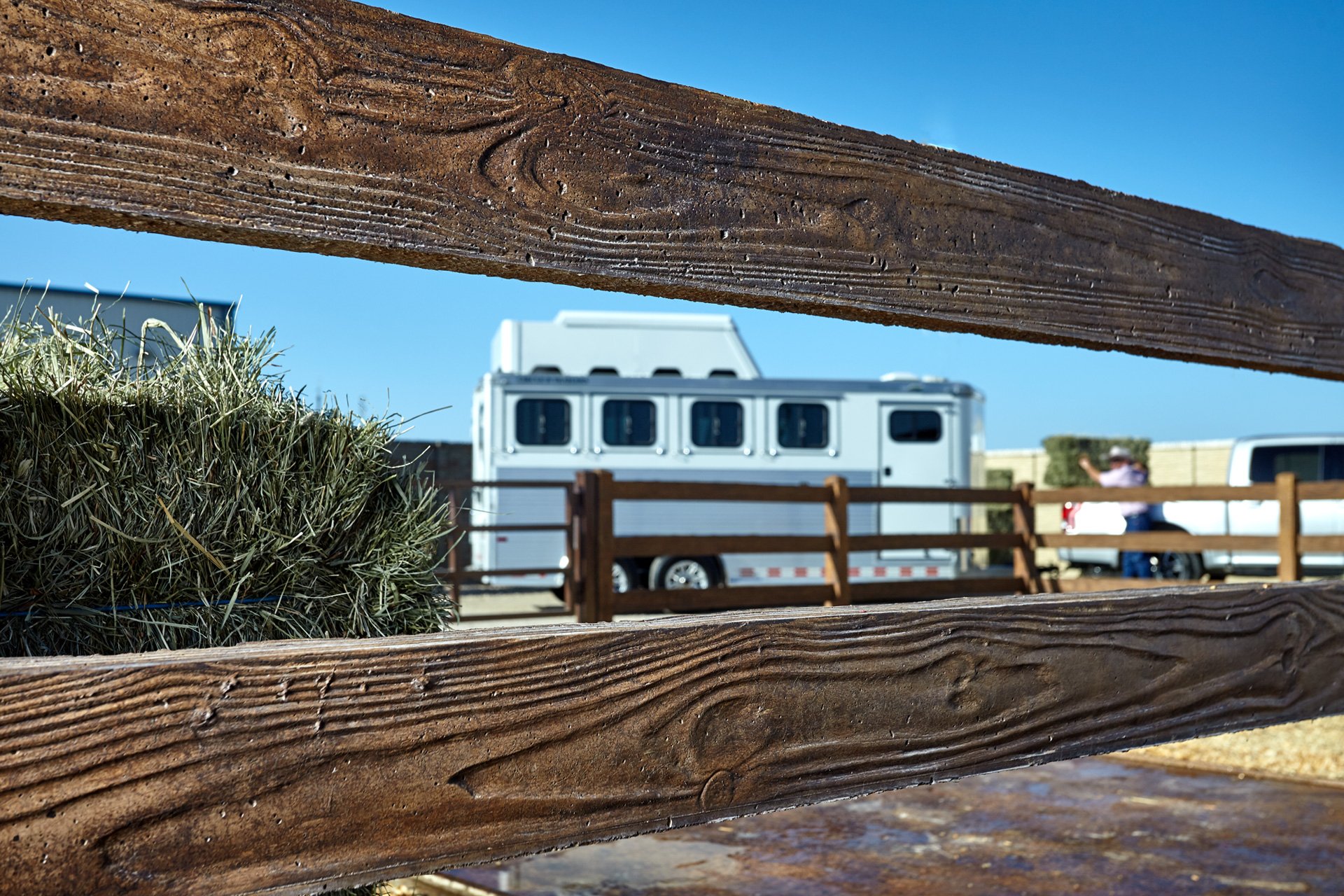 A wooden fence with horizontal slats, a bundle of dry grass, and a white horse trailer in the background, with a person taking a photo and a vehicle nearby under a clear blue sky.