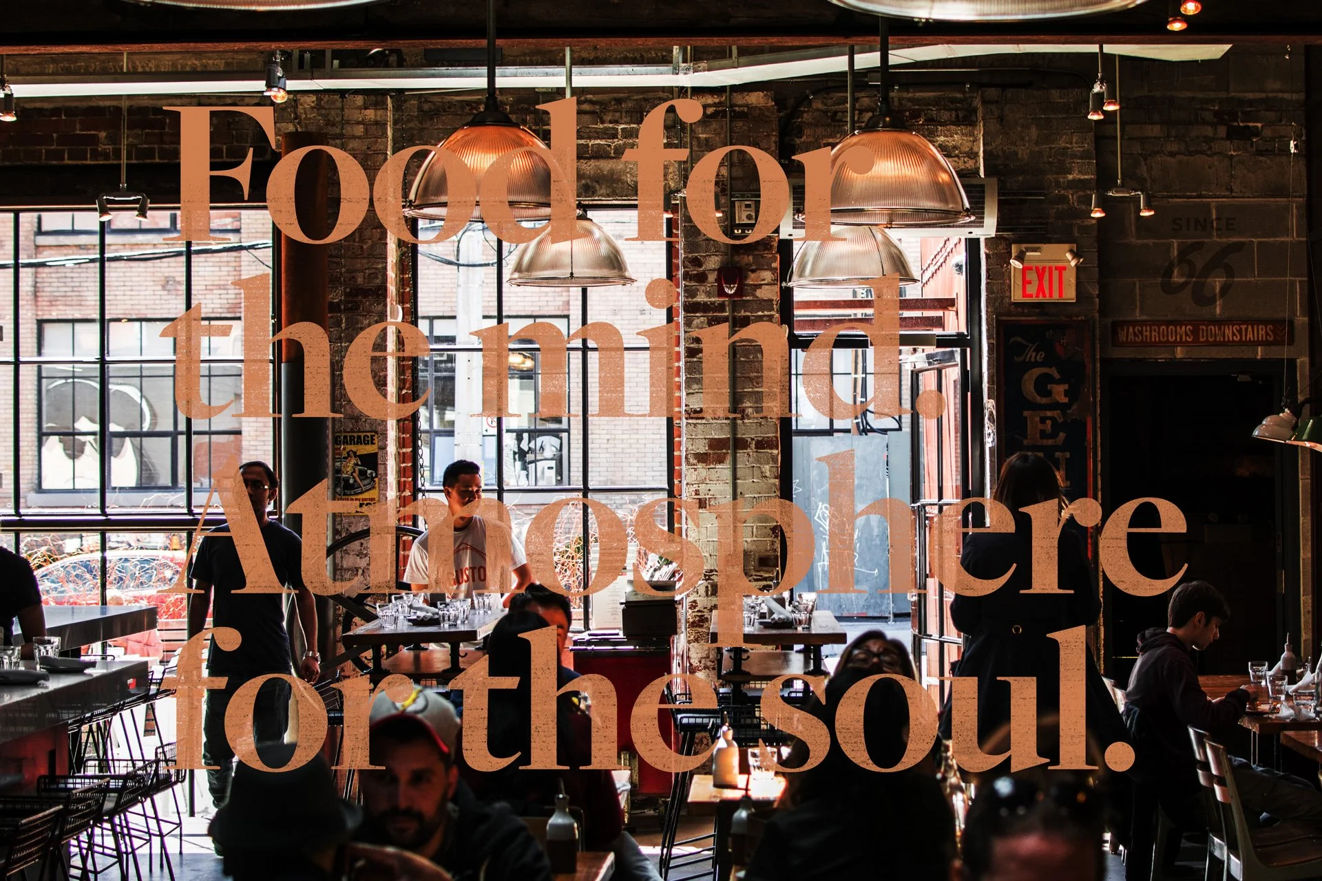 Inside a lively restaurant with large windows, hanging pendant lights, and exposed brick walls. Overlaid text reads: 'Food for the mind, atmosphere for the soul.'